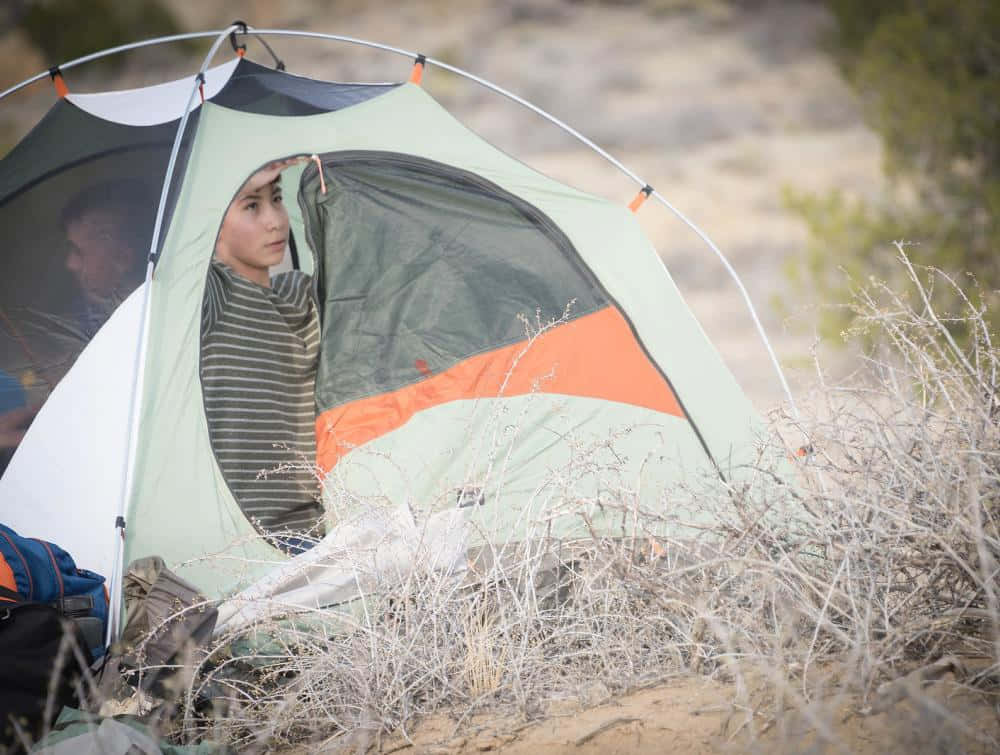 Fall Camping 桌布 壁紙 — 一個穿著條紋襯衫的人從一個藏在沙漠乾燥灌木叢中的綠色和橙色帳篷裡探出頭來。