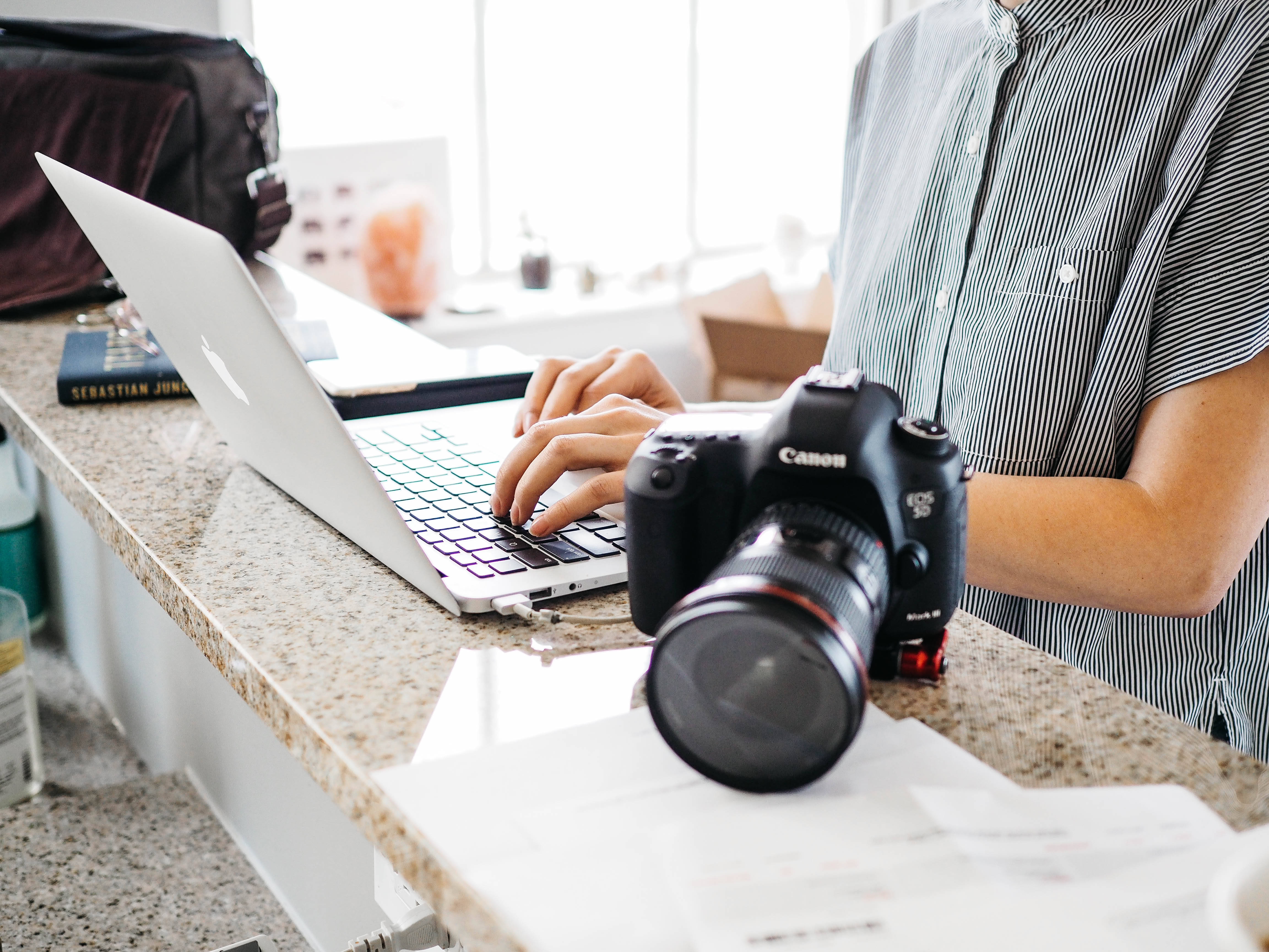 typing 桌布 壁紙 — 一個穿著條紋襯衫的人正在花崗岩檯面上的Canon DSLR相機旁邊的筆記型電腦上打字。