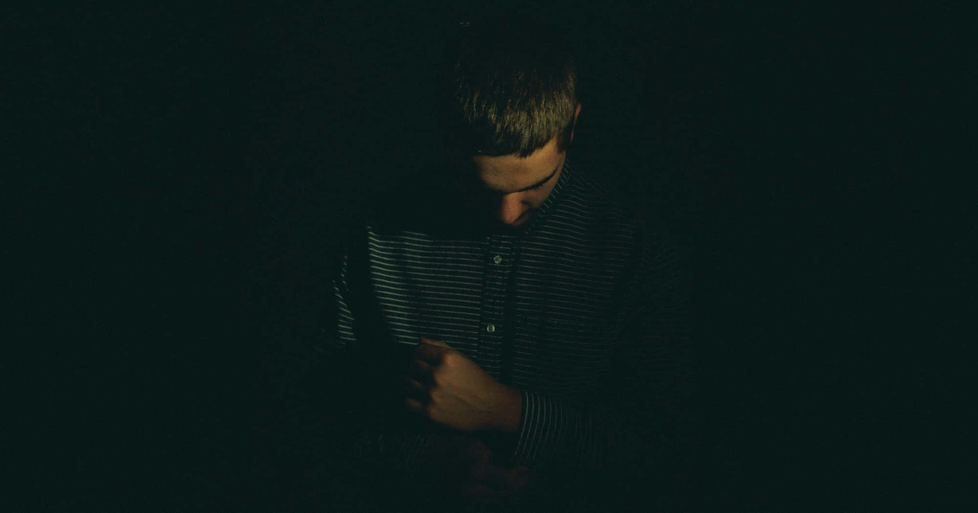 Depressed 桌布 壁紙 — 一個穿著條紋襯衫的男人站在近乎黑暗的環境中，低著頭，手放在胸前，被一道聚焦的光線照亮。