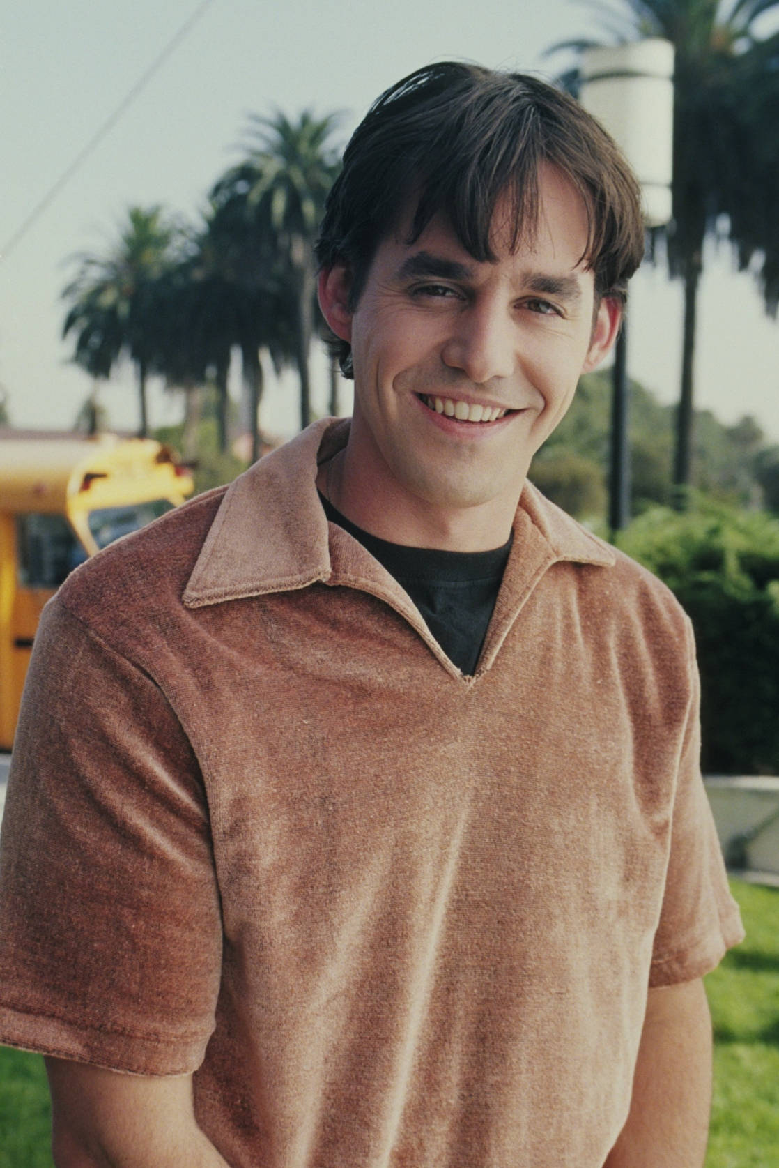 Nicholas Brendon 桌布 壁紙 — 一個穿著棕色Polo衫的開朗男子在戶外微笑，背景有棕櫚樹和黃色巴士。