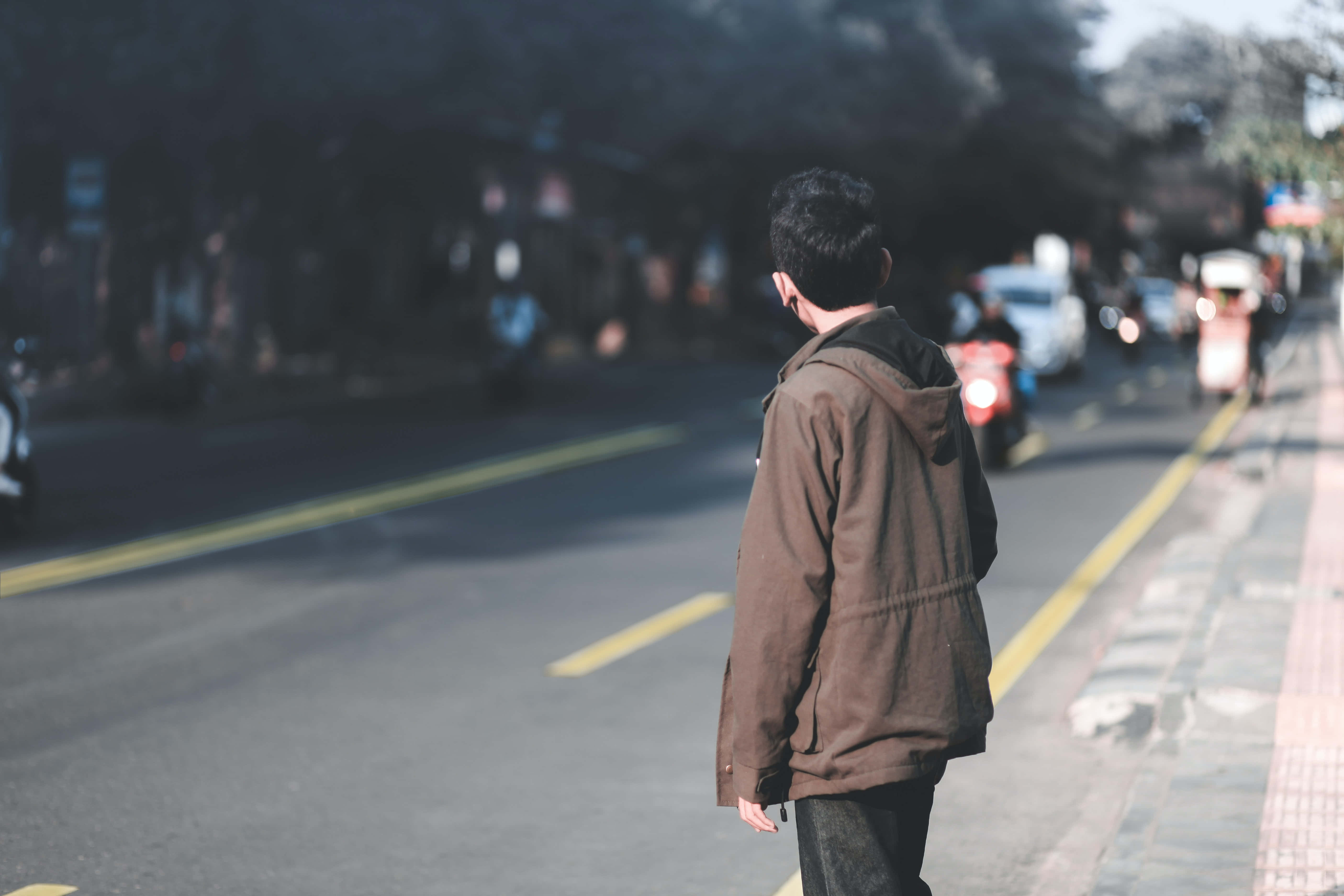 Waiting 桌布 壁紙 — 一個穿著棕色外套的人獨自站在陽光普照的都市街道上，機車和汽車駛過。