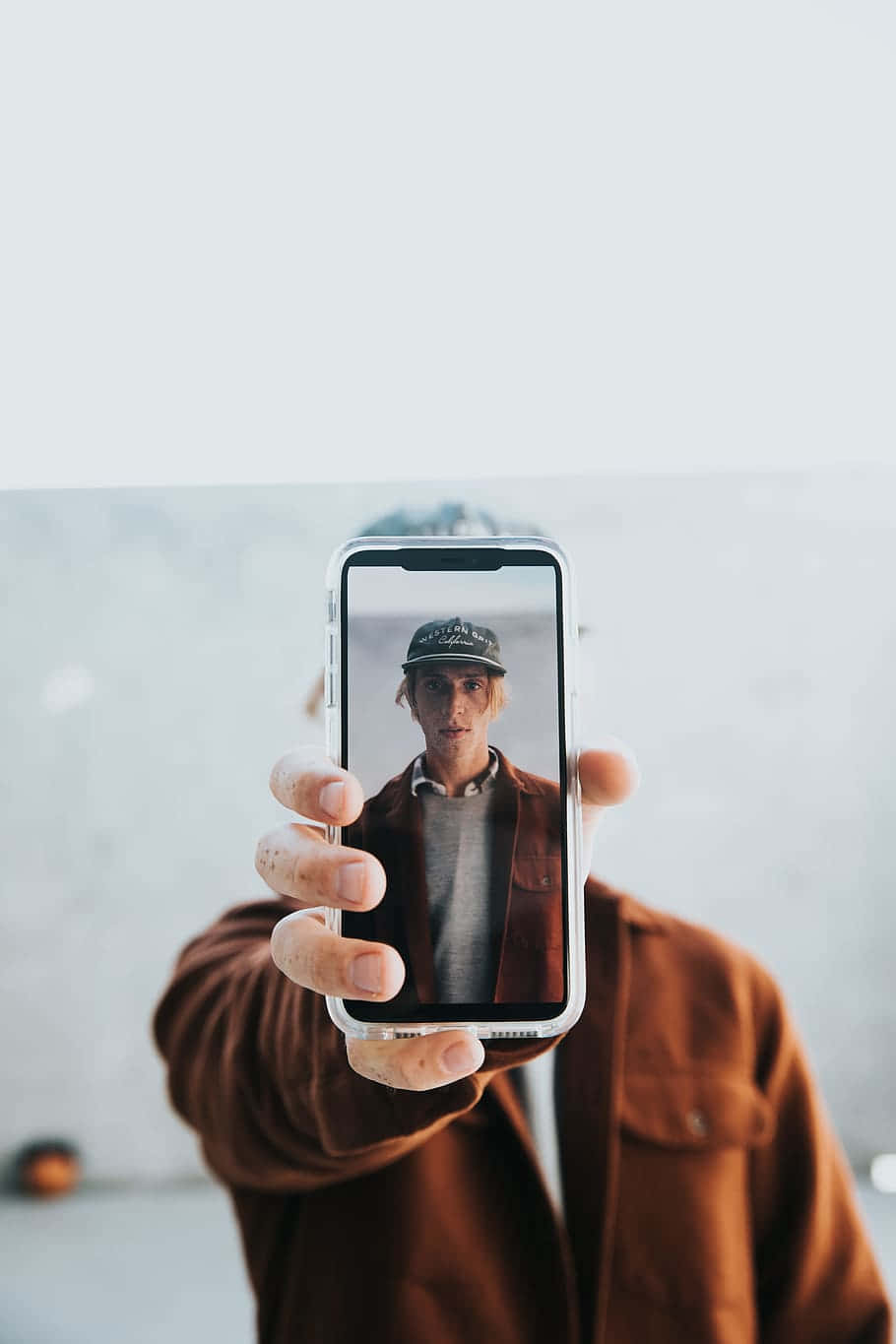iPhone Xs Max Apple 桌布 壁紙 — 一個穿著棕色外套的人舉起手機，顯示自己戴著帽子的臉，背景模糊。