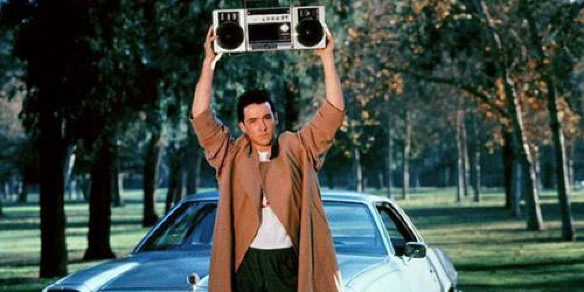 John Cusack 桌布 壁紙 — 一個穿著棕色外套的男人在陽光明媚的公園裡，旁邊有一輛銀色汽車，他勝利地將一個手提音響舉過頭頂。