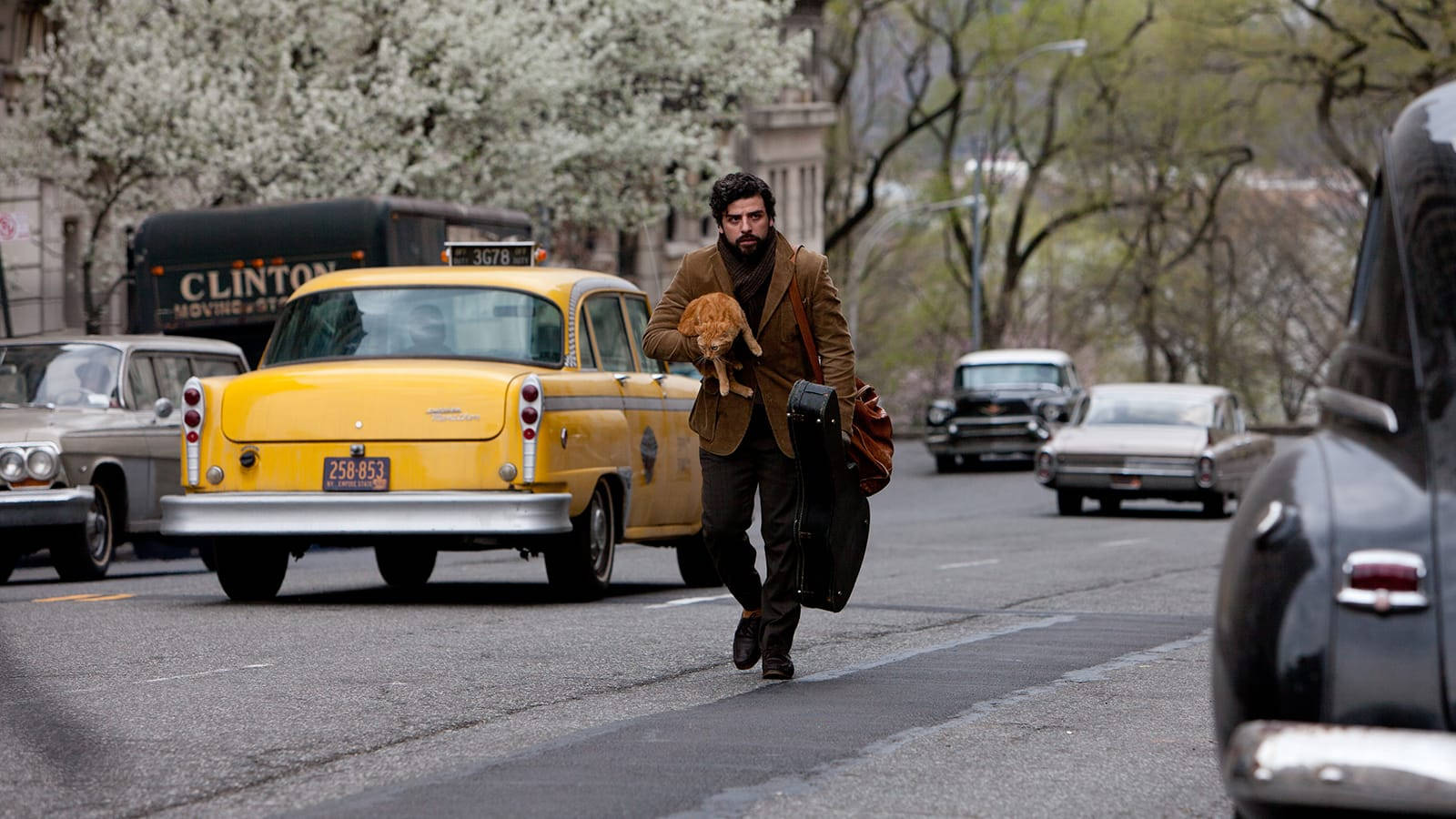 inside llewyn davis 桌布 壁紙 — 一個穿著棕色外套的男人抱著一隻貓和一個吉他盒，跑過一條繁忙的街道，背景有復古黃色計程車和開花的樹木。