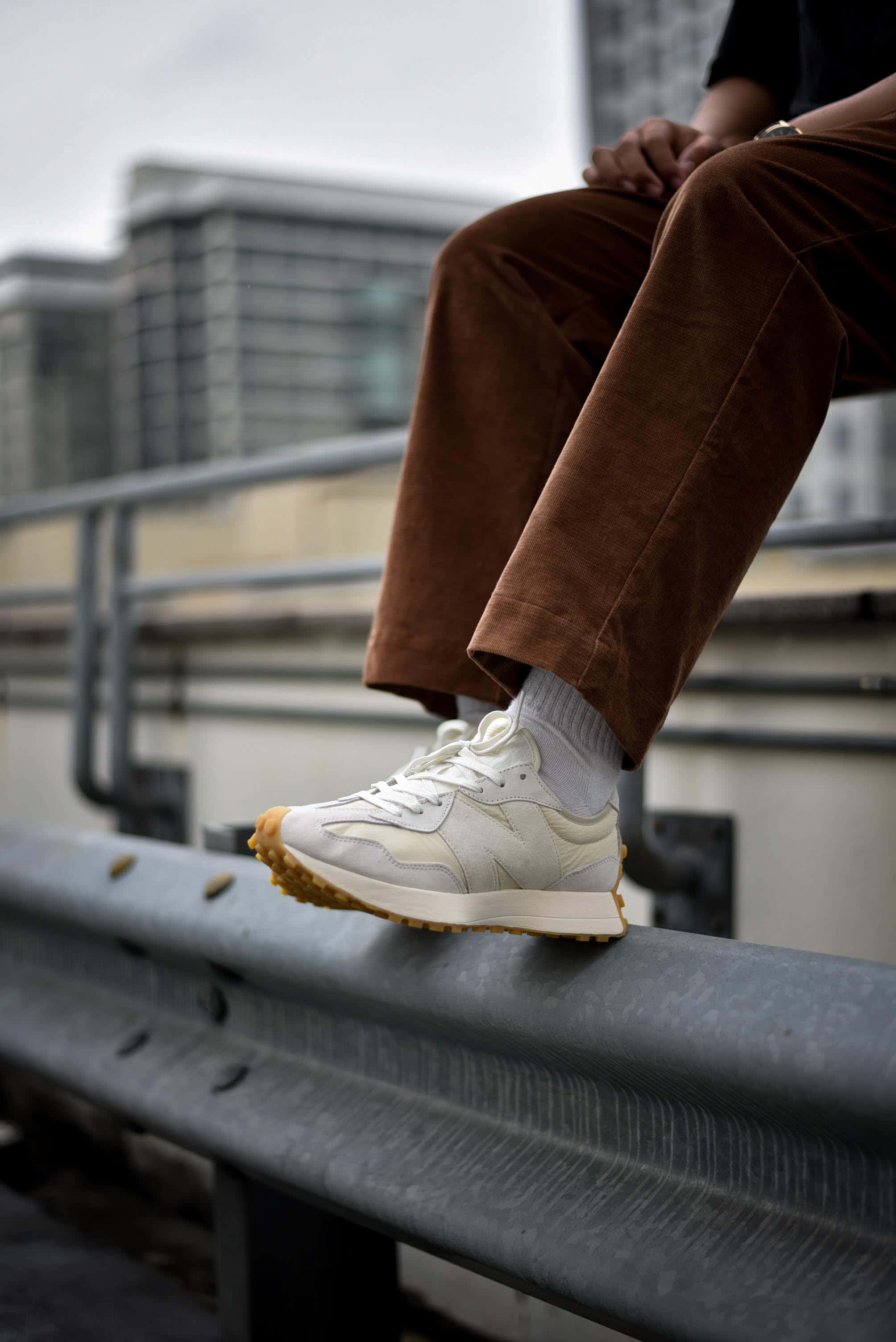 New Balance 桌布 壁紙 — 一個穿著棕色燈芯絨褲子和白色運動鞋的人，在模糊的城市景觀背景下，隨意地靠在金屬欄杆上休息。