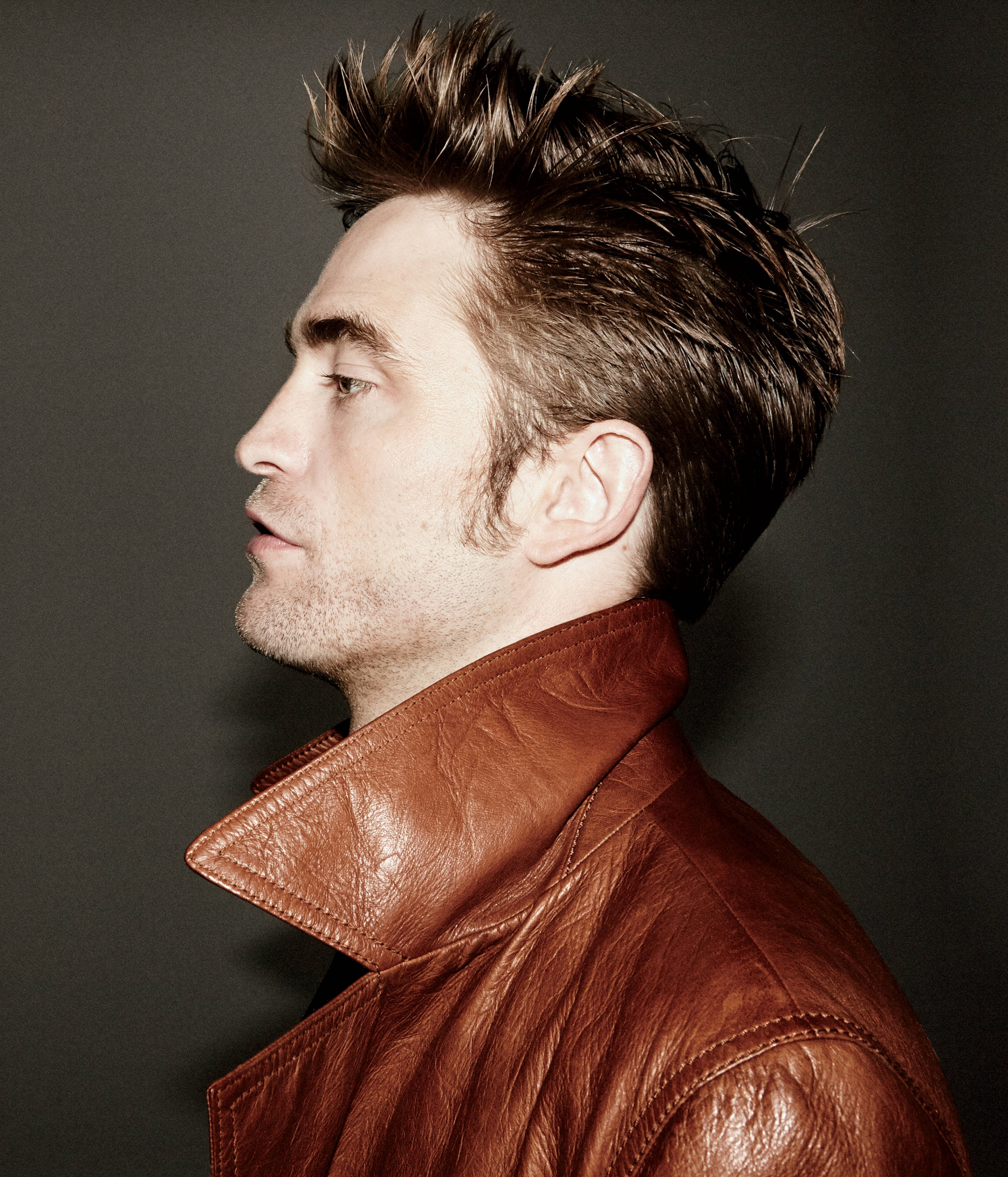 Robert Pattinson 桌布 壁紙 — 一個穿著棕色皮夾克的男人，在柔和的灰色背景下沉思地凝視。