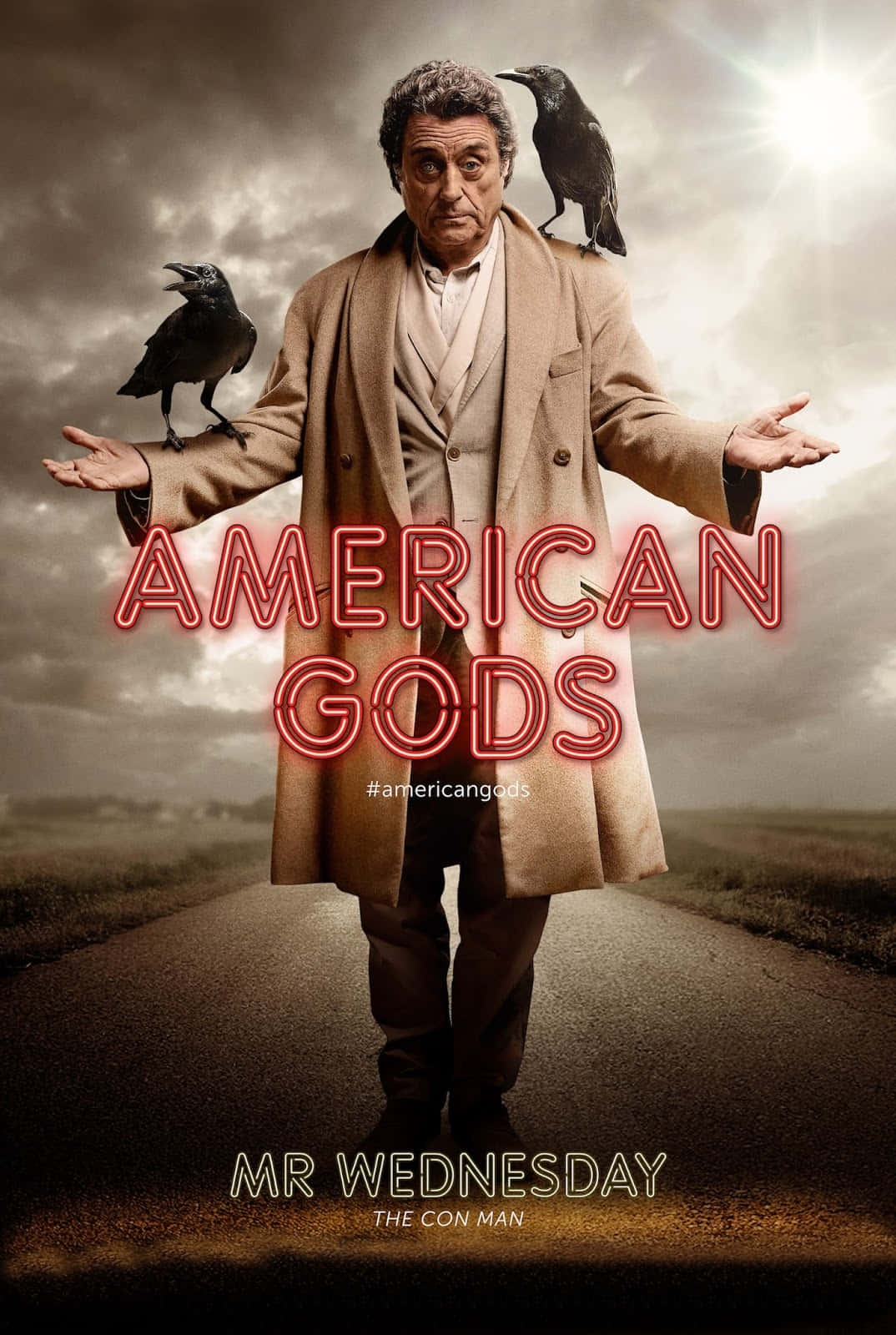 American Gods 桌布 壁紙 — 一個穿著棕褐色外套的男人站在荒涼的道路上，兩隻烏鴉棲息在他的手臂上，天空多雲，戲劇性的陽光穿透雲層。