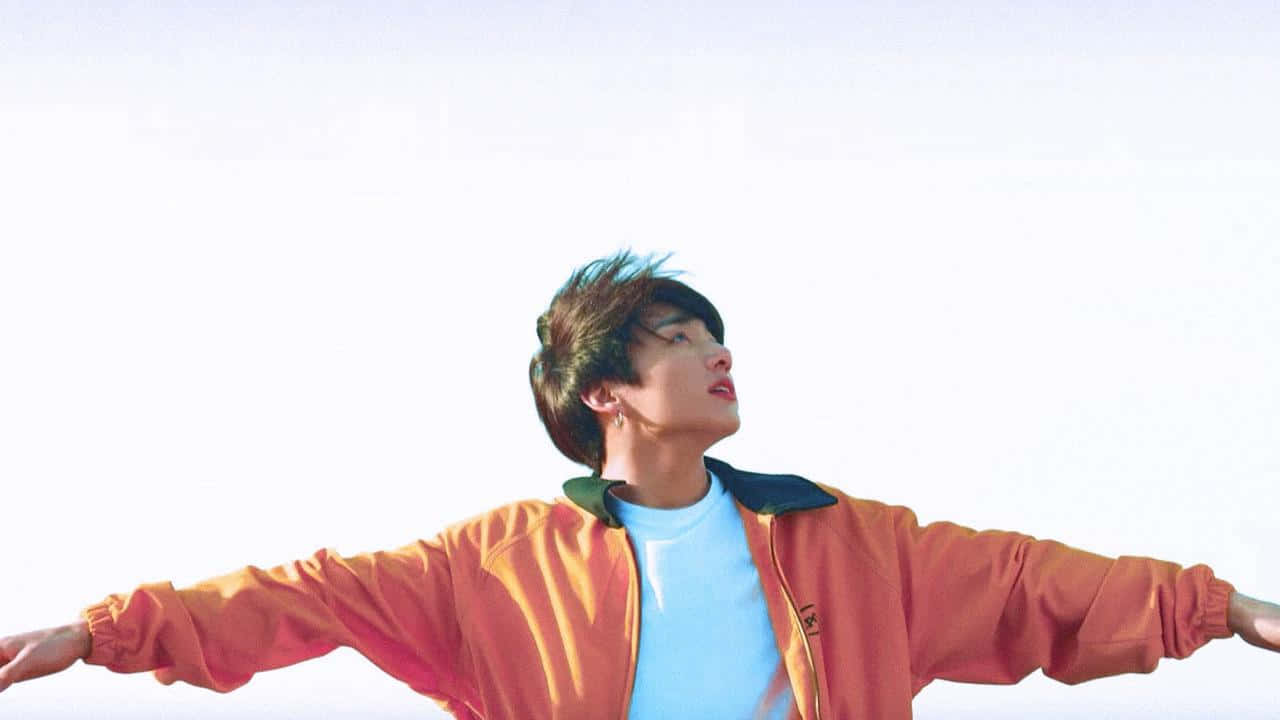 Jungkook 桌布 壁紙 — 一個穿著橙色外套的年輕人張開雙臂，面向明亮的白色天空，頭部向上傾斜，感受自由。