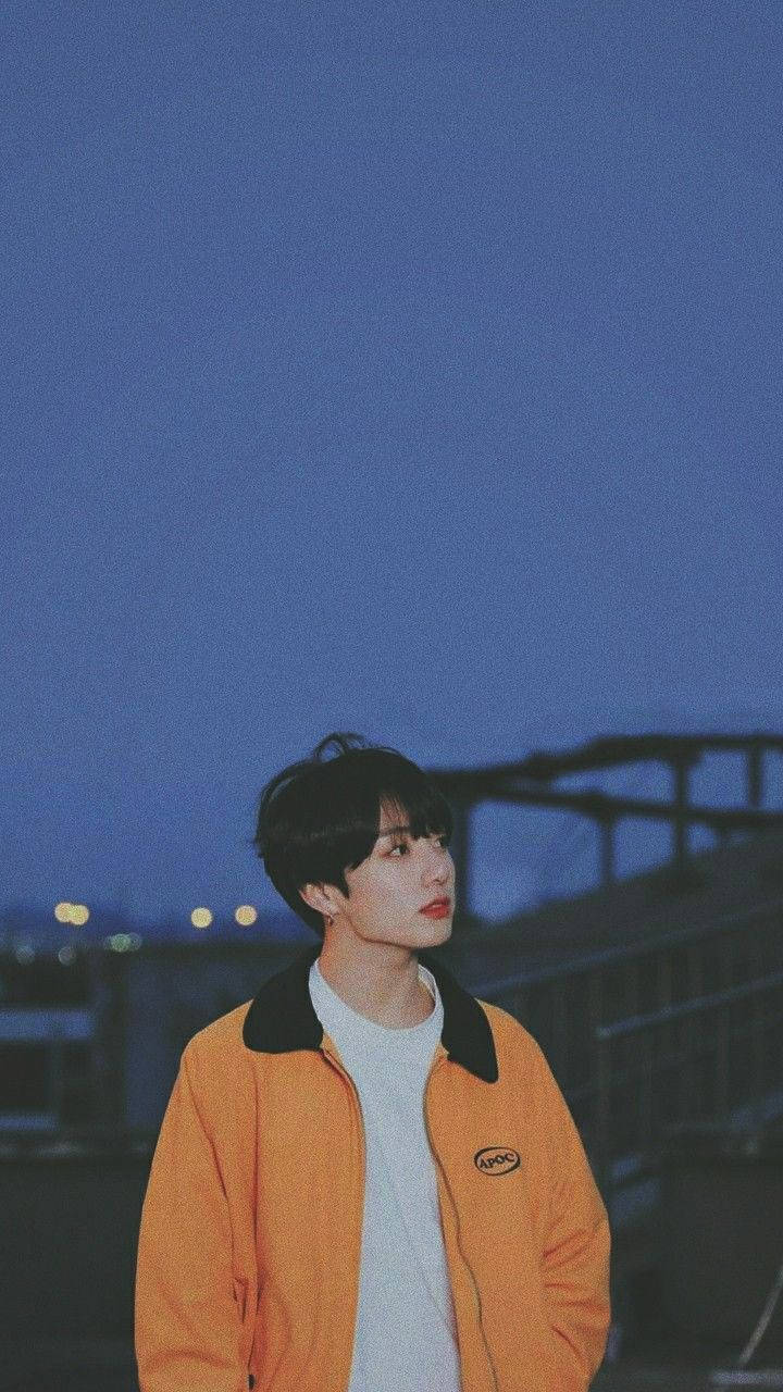 Jungkook 桌布 壁紙 — 一個穿著橙色外套的年輕男子站在暮色天空下，遠處的城市燈光在他身後柔和地閃爍。