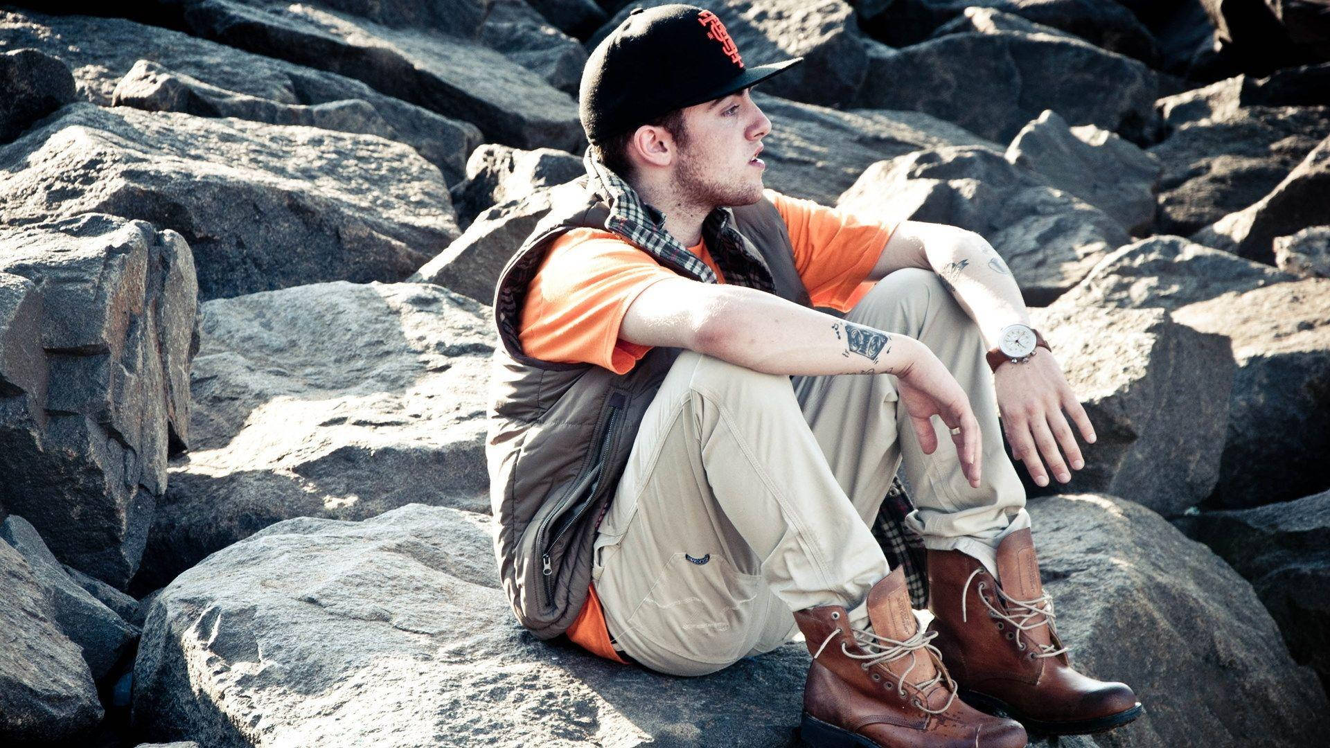 Mac Miller 桌布 壁紙 — 一個穿著橙色襯衫和帽子的年輕男子盤腿坐在陽光普照的灰色岩石上，若有所思地凝視遠方。