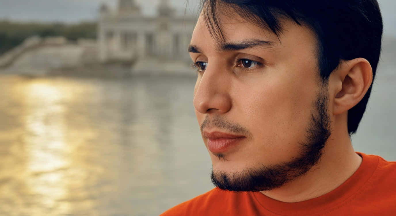 José Rafael Cordero Sánchez 壁紙 — 一個穿著橙色襯衫的男人若有所思地凝視著一座模糊的歷史建築，倒映在金色的水中。