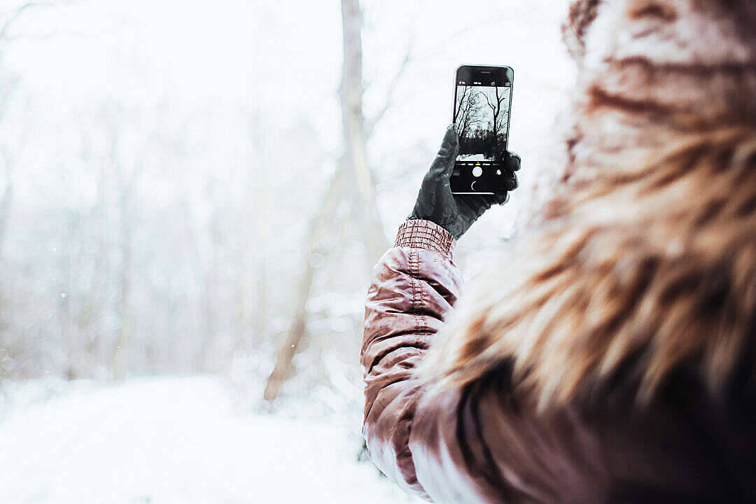 photography iPhone 桌布 壁紙 — 一個穿著毛皮大衣和手套的人正在冬天用手機拍攝雪覆蓋的森林場景。