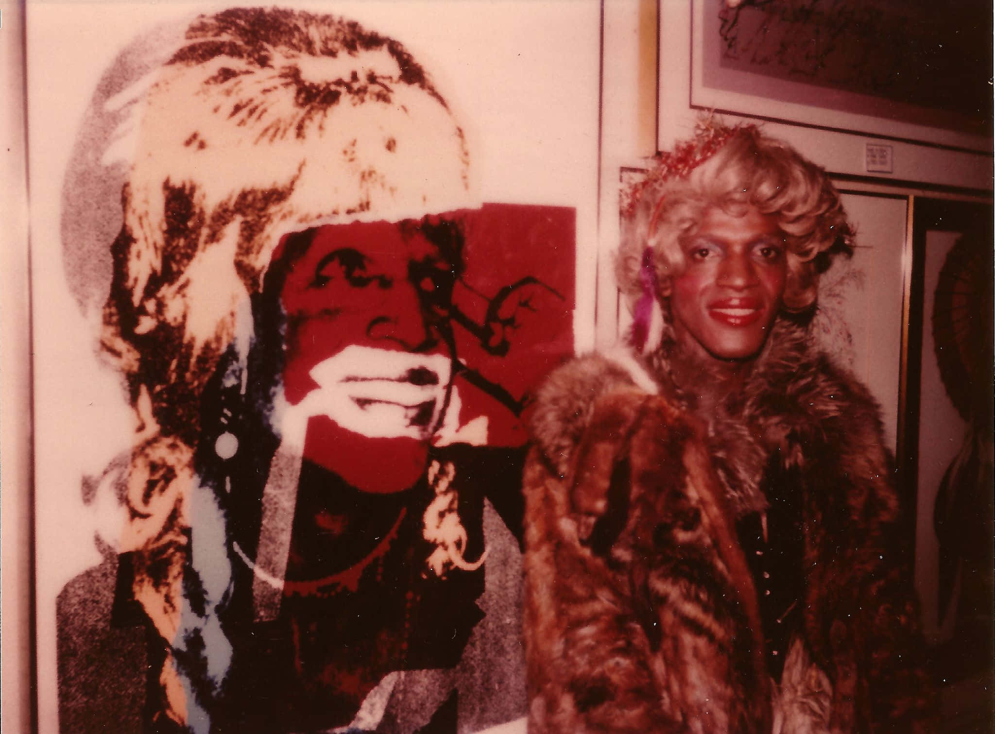 andy warhol 桌布 壁紙 — 一個穿著毛皮大衣和捲曲假髮的人站在一個大型普普藝術肖像旁邊，肖像中的男人有紅色的嘴巴和白髮。
