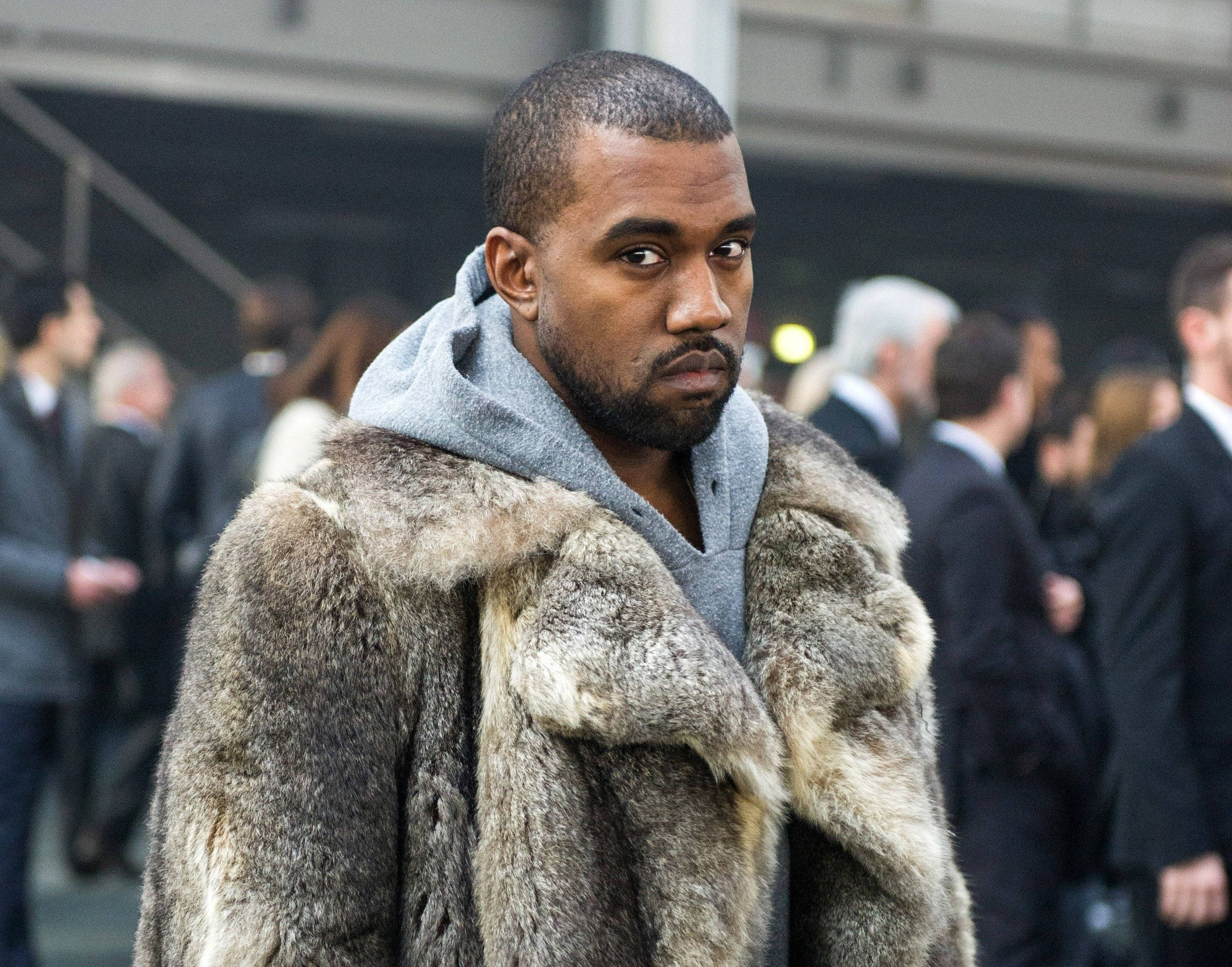 Kanye West 桌布 壁紙 — 一個穿著毛皮大衣和連帽衫的男人在模糊的人群中凝視著相機，身處城市環境。