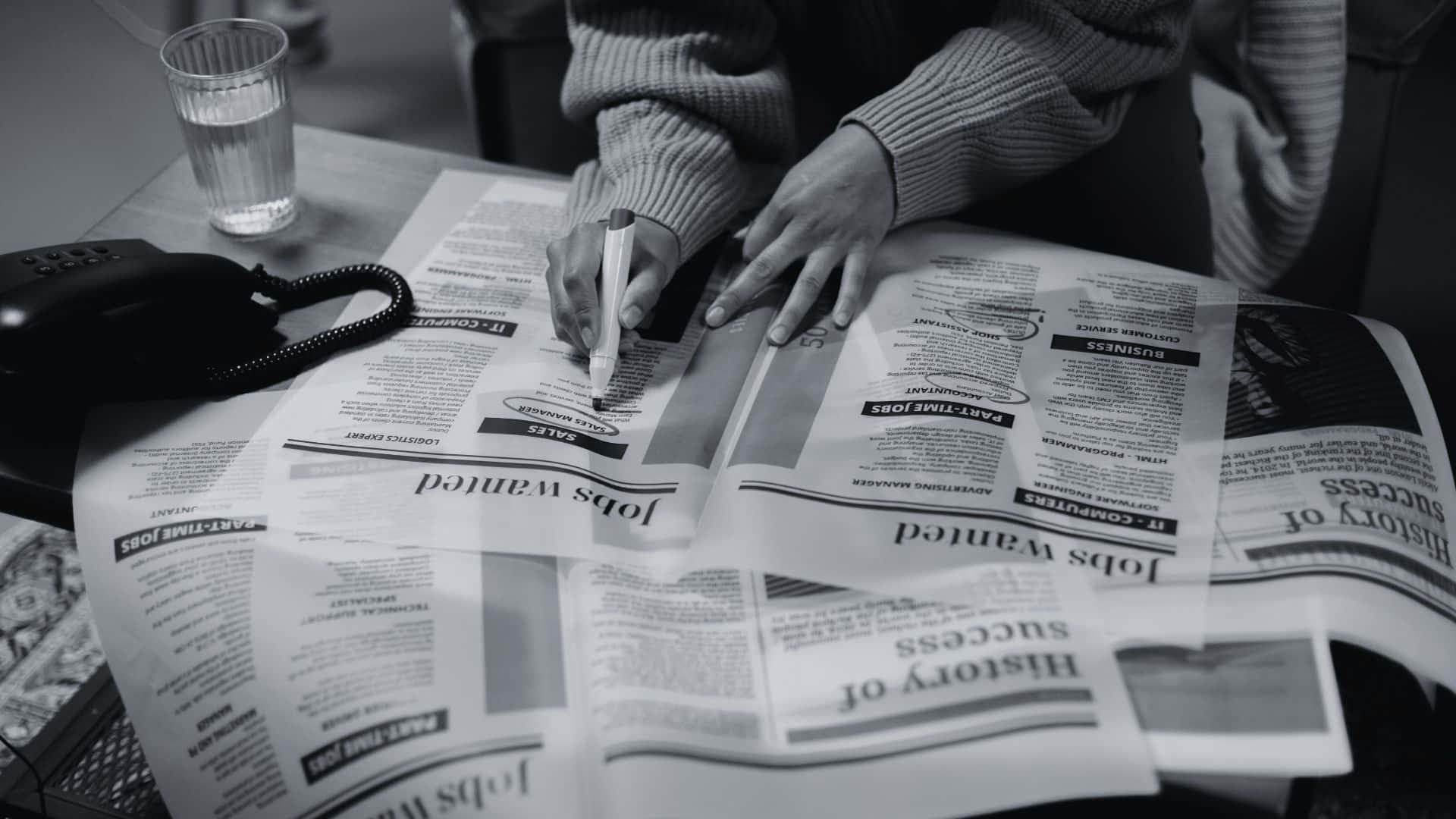 Black And White Newspaper 桌布 壁紙 — 一個穿著毛衣的人正在一份刊登招聘廣告的報紙上寫字，旁邊放著一杯水和一支電話。
