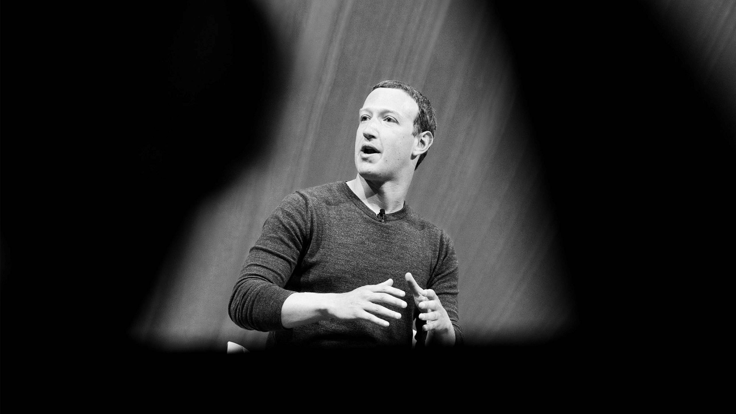 Zuckerberg 桌布 壁紙 — 一個穿著毛衣的男人在聚光燈下熱情地說話，周圍是黑暗的剪影。