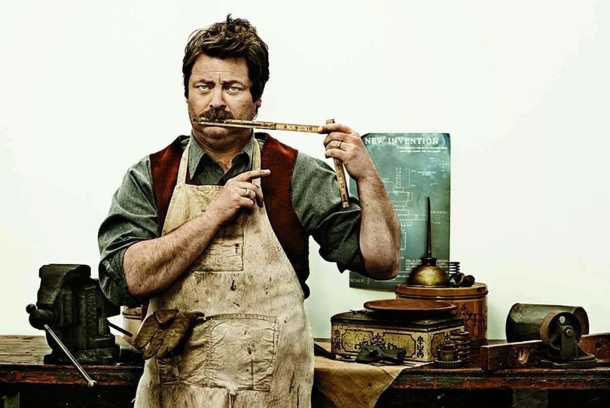 nick offerman 桌布 壁紙 — 一個穿著沾污圍裙的男人在一個雜亂的工作室裡，將尺子放在鬍鬚上，周圍滿是工具和藍圖。