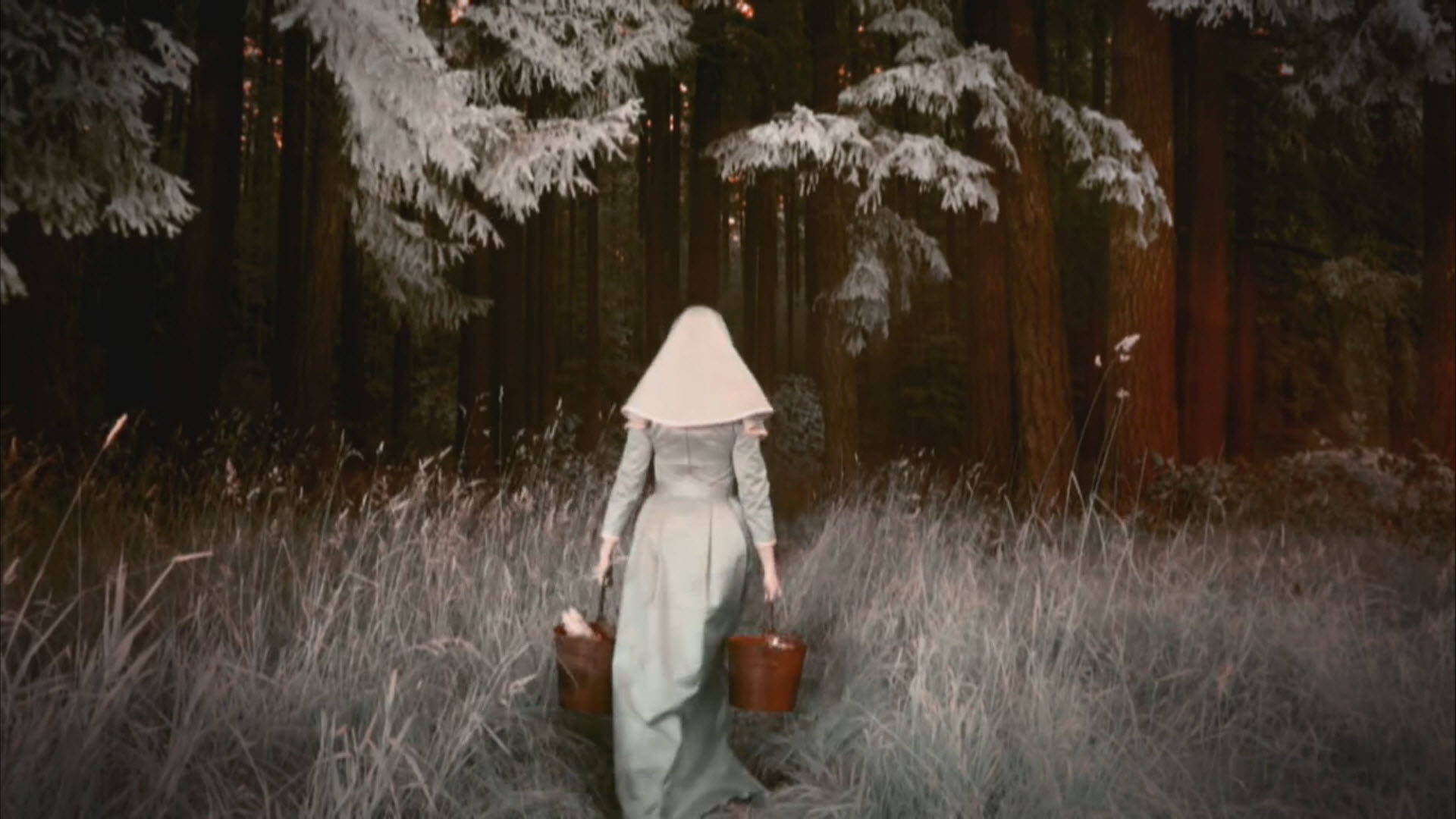 American Horror Story 桌布 壁紙 — 一個穿著淡色長袍和錐形頭飾的人物，穿過雪地森林，提著兩個水桶。