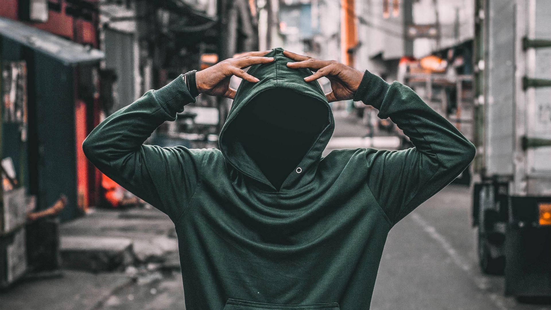 City Street 桌布 壁紙 — 一個穿著深綠色連帽衫的人在一條狹窄的城市小巷中將帽子拉過臉部。