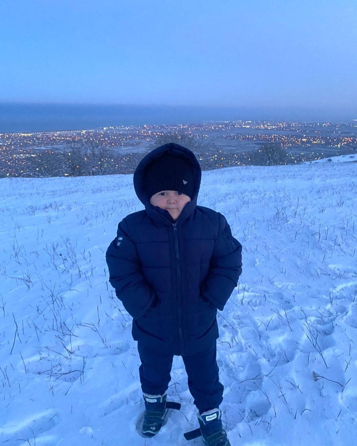 Winter 桌布 壁紙 — 一個穿著深色冬季外套的小孩站在雪丘上，俯瞰著黃昏時分燈火通明的城市。