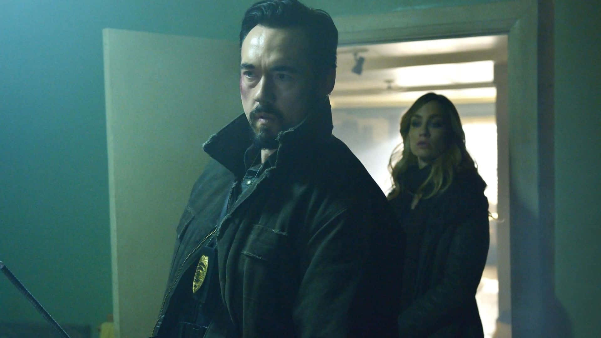 Kevin Durand 桌布 壁紙 — 一個穿著深色外套、帶有徽章的男人站在燈光昏暗的房間裡，一個女人從他身後探頭張望。