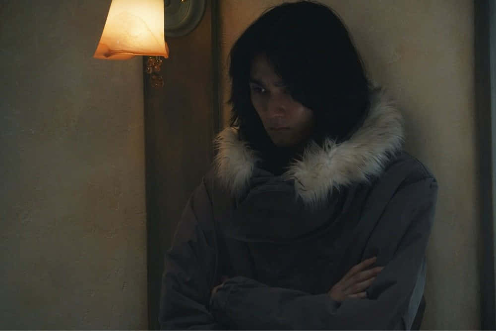 Tokyo Ghoul Movie 桌布 壁紙 — 一個穿著深色外套、白色毛領的人，在昏暗的房間裡，在壁燈的溫暖光芒下交叉雙臂站立。