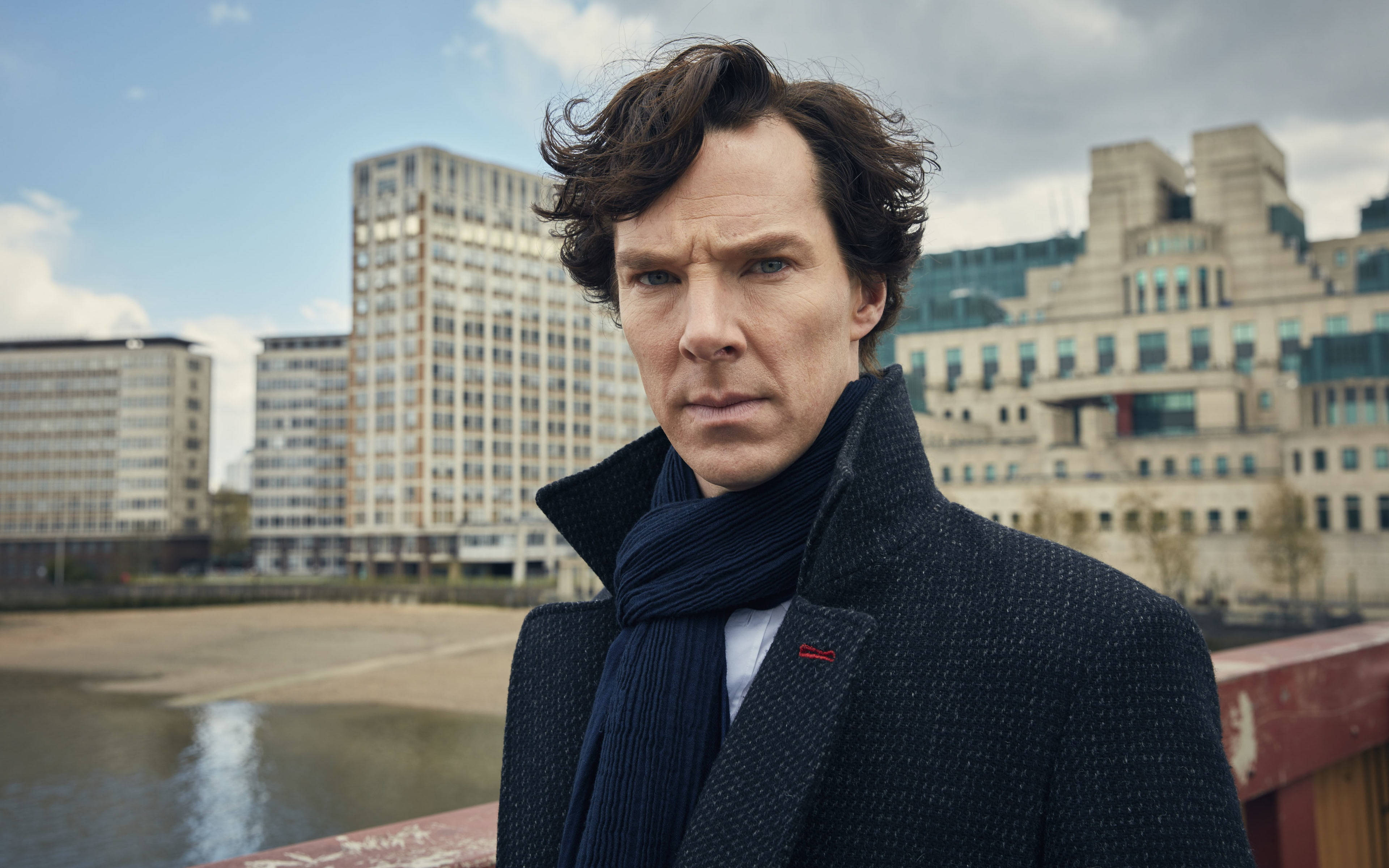 Sherlock Holmes 桌布 壁紙 — 一個穿著深色外套和圍巾的男人站在橋上，俯瞰著一條河流，身後是現代城市建築，天空多雲。