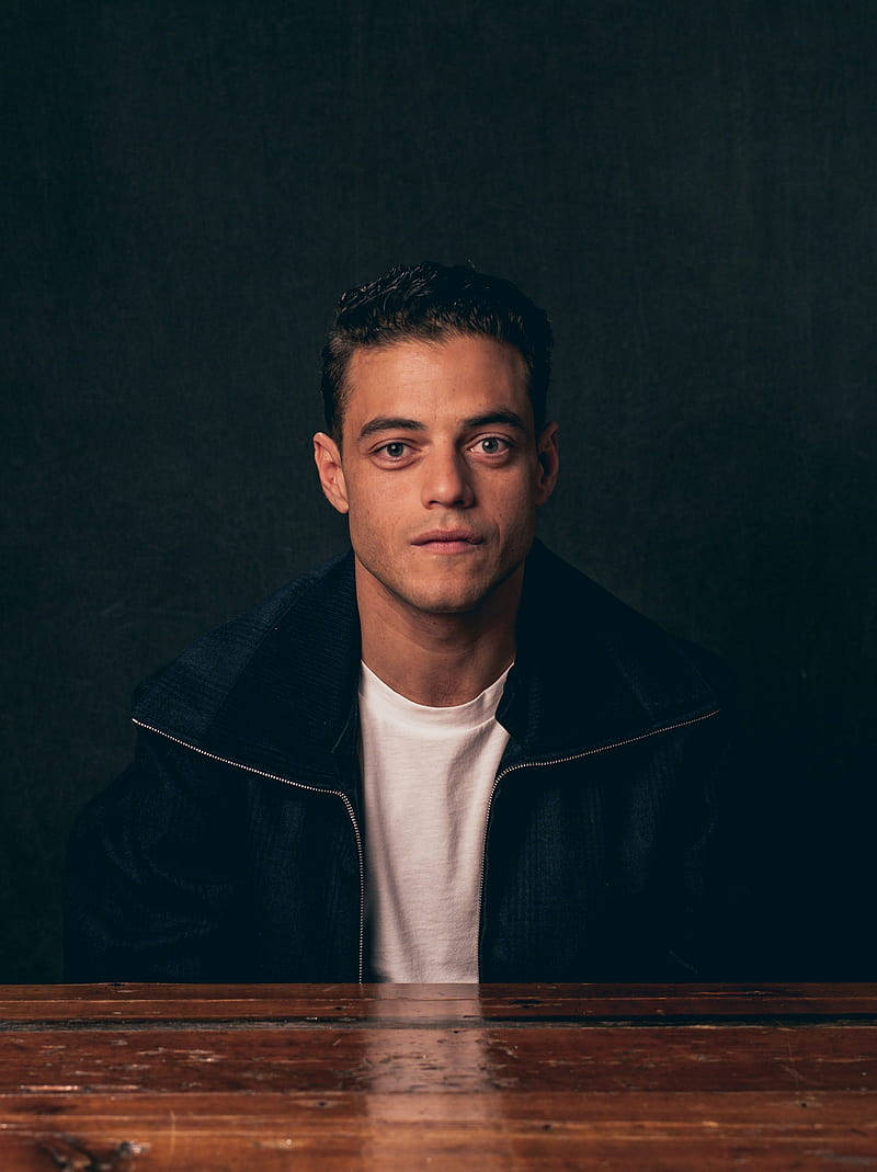 Rami Malek 桌布 壁紙 — 一個穿著深色外套和白色T恤的年輕男子坐在木桌旁，背景是陰暗的氛圍。