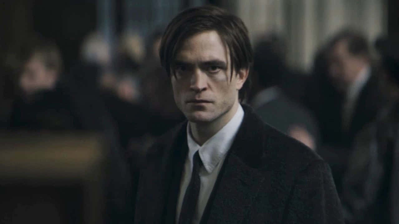 Bruce Wayne 桌布 壁紙 — 一個穿著深色外套和領帶的男人在燈光昏暗、擠滿人的房間裡，身後有模糊的人影，專注地向前凝視。