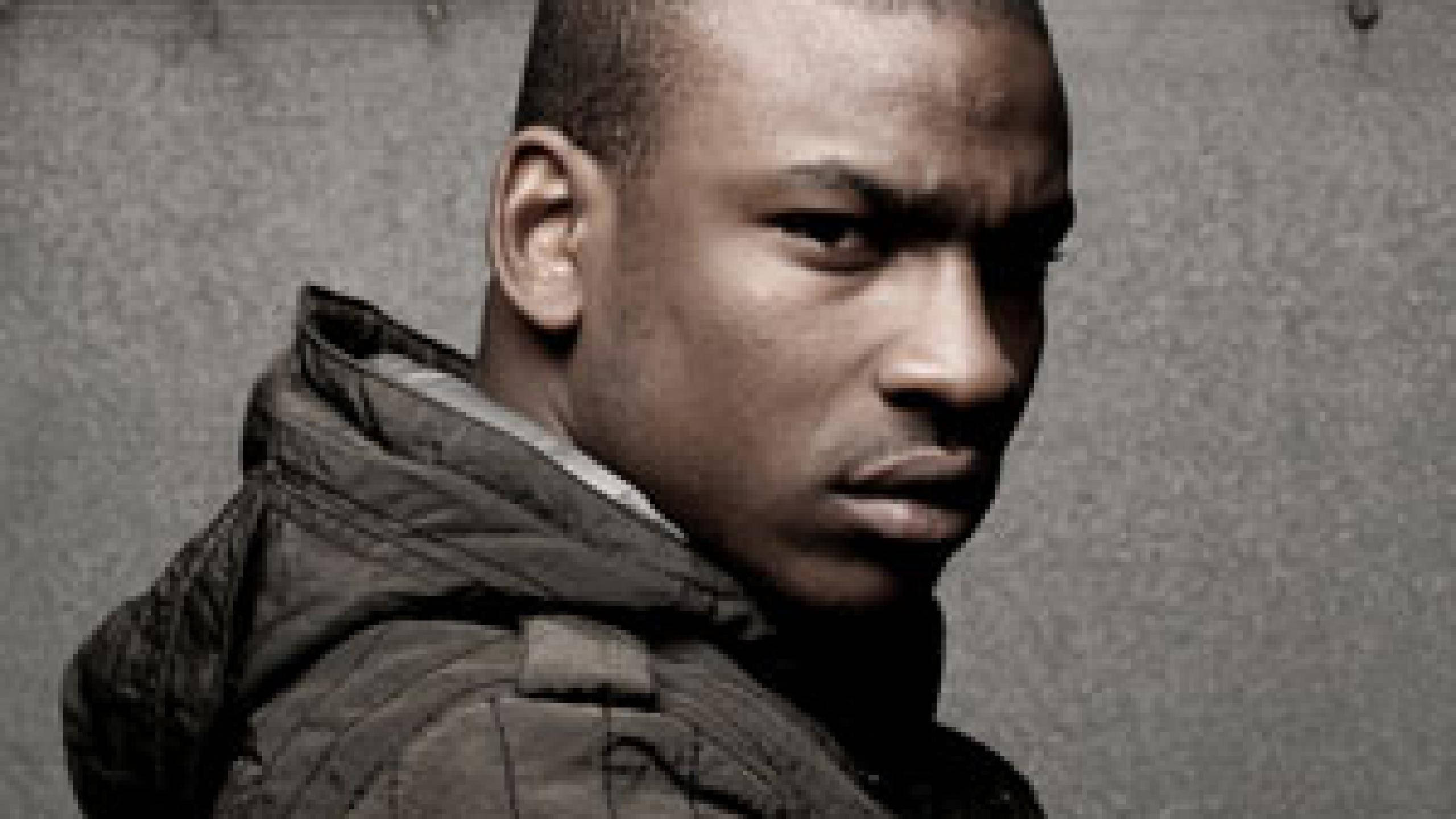 Skepta 桌布 壁紙 — 一個穿著深色外套的嚴肅年輕男子，在單色調中對著粗糙有紋理的牆壁凝視。