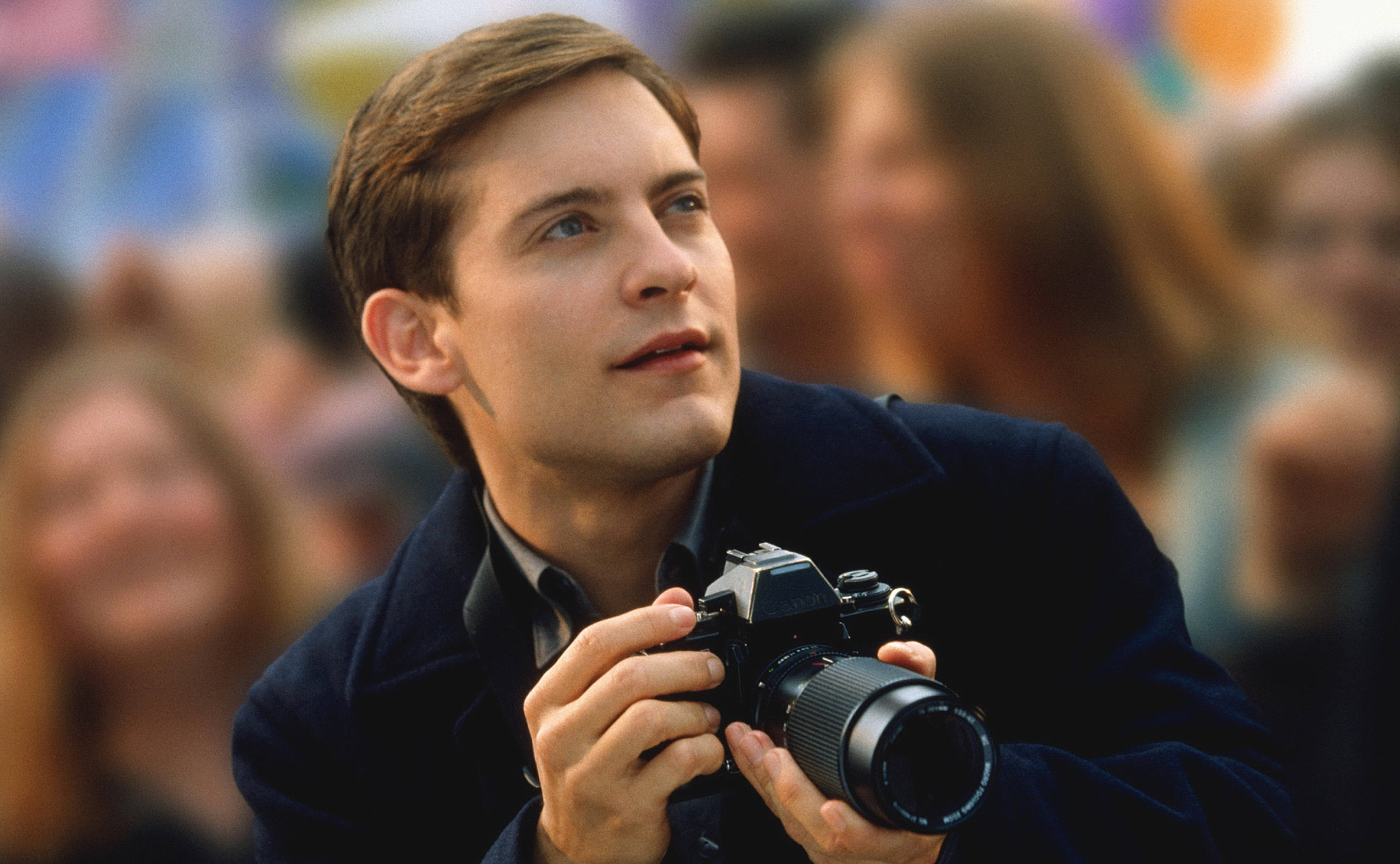 Tobey Maguire 桌布 壁紙 — 一個穿著深色外套的年輕男子在熙攘的人群中手持復古相機，帶著專注的好奇心向上凝視。