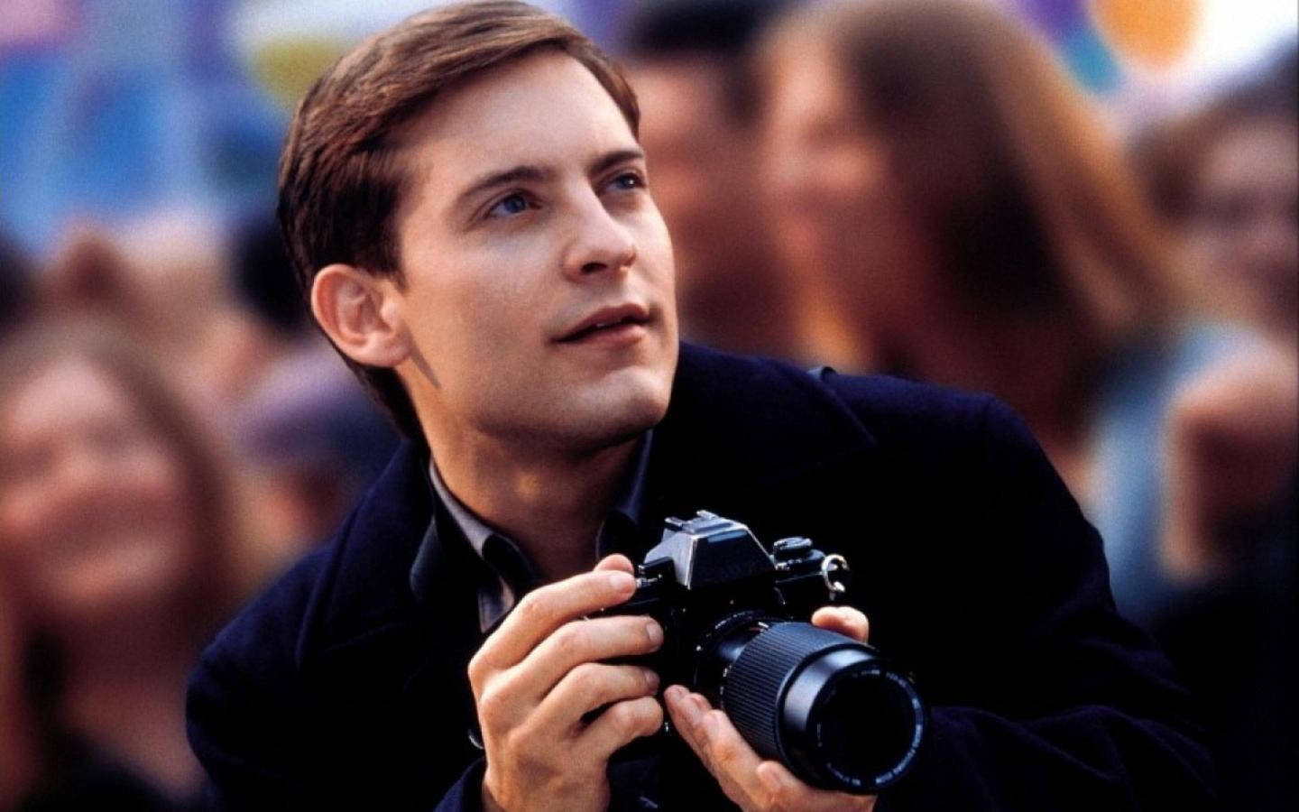Tobey Maguire 桌布 壁紙 — 一個穿著深色外套的年輕男子拿著相機，在模糊的人群背景下專注地向上凝視。