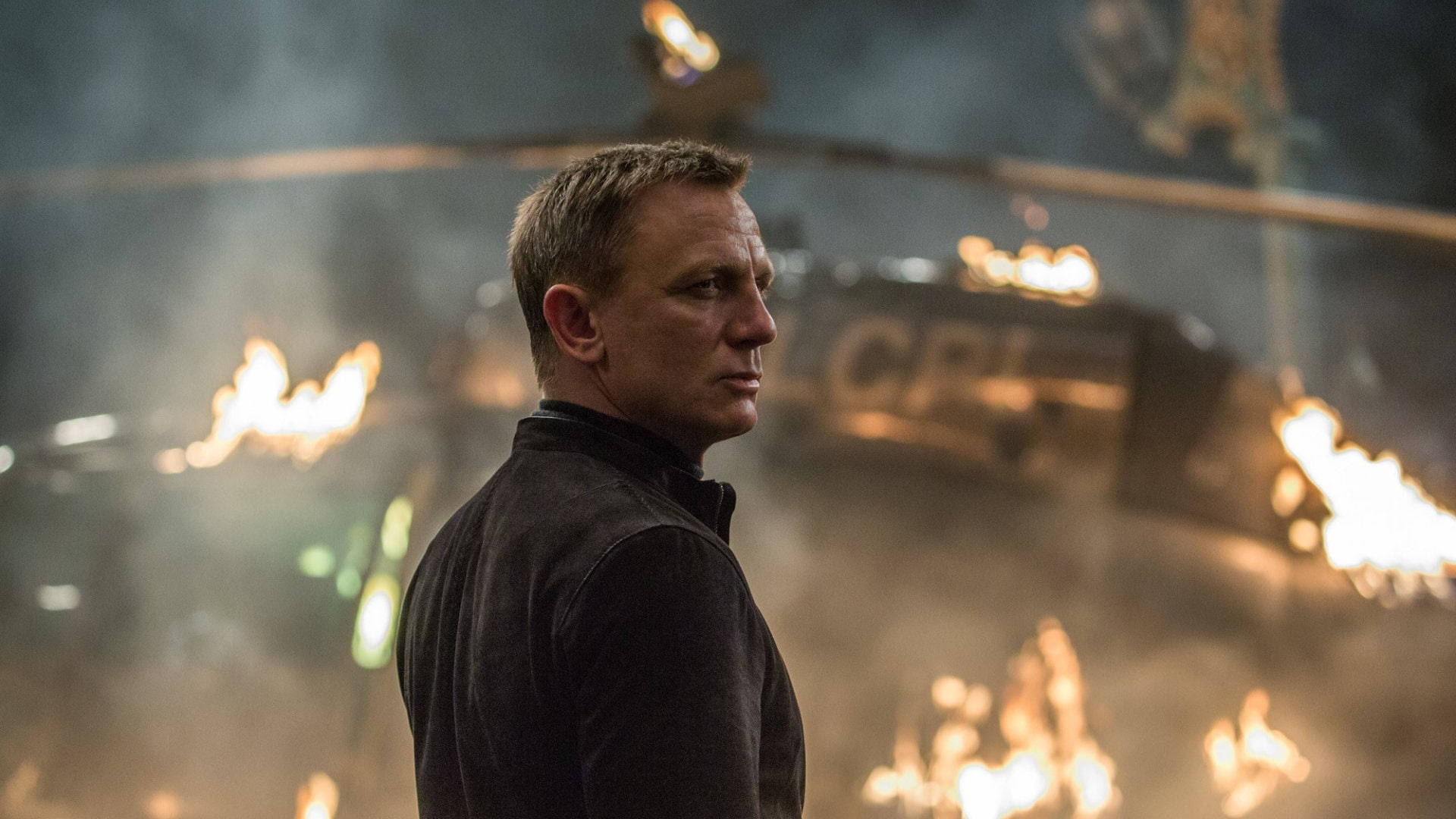 Daniel Craig 桌布 壁紙 — 一個穿著深色外套的男人，在熾熱爆炸和煙霧的背景下堅定地站立。