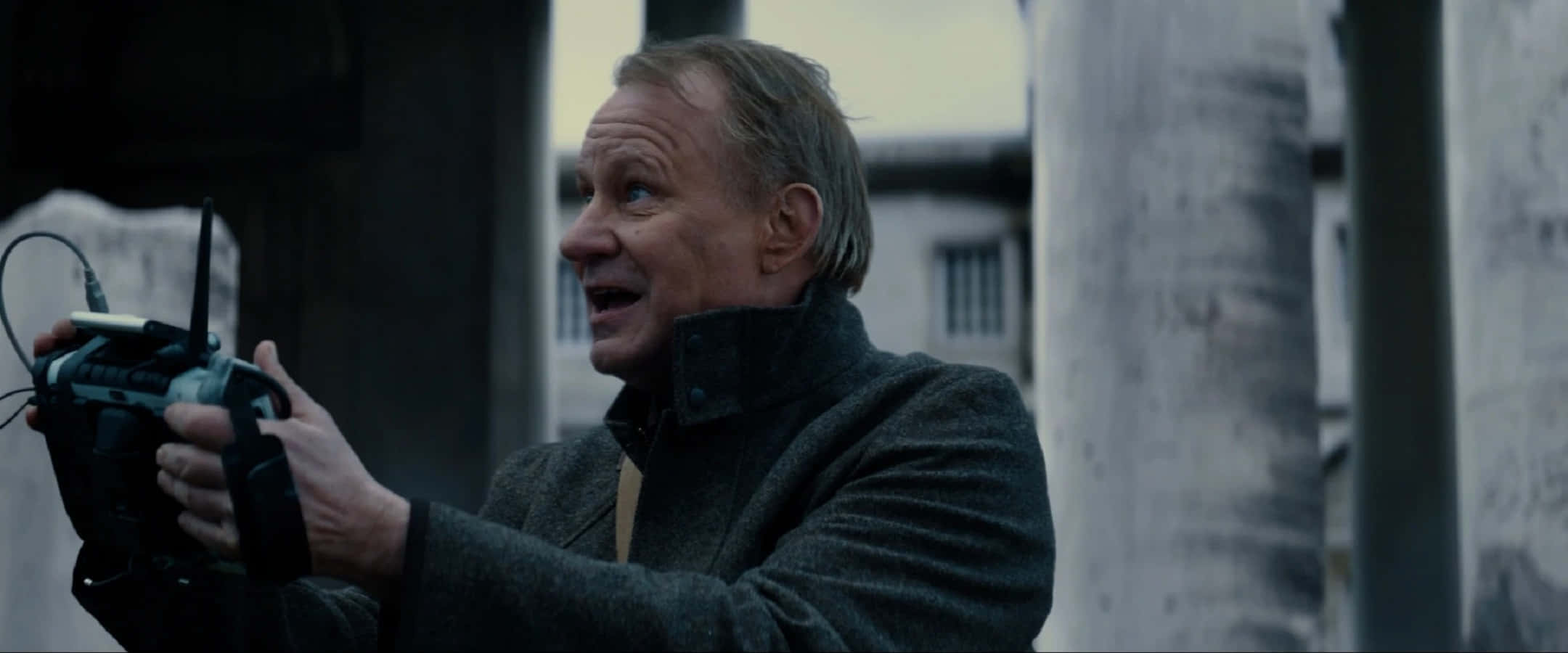 Erik Selvig 桌布 壁紙 — 一個穿著深色外套的男人，在粗糙的城市背景下，帶著強烈的表情拿著無線電發射器。