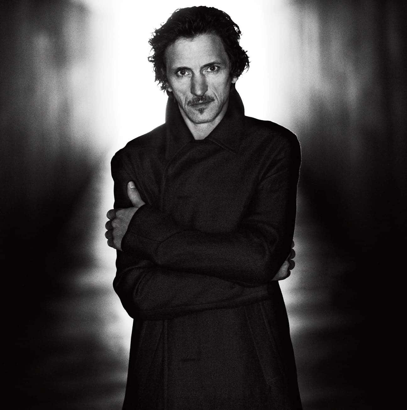 John Hawkes 桌布 壁紙 — 一個穿著深色外套的男人交叉雙臂站在模糊的高對比黑白背景前。