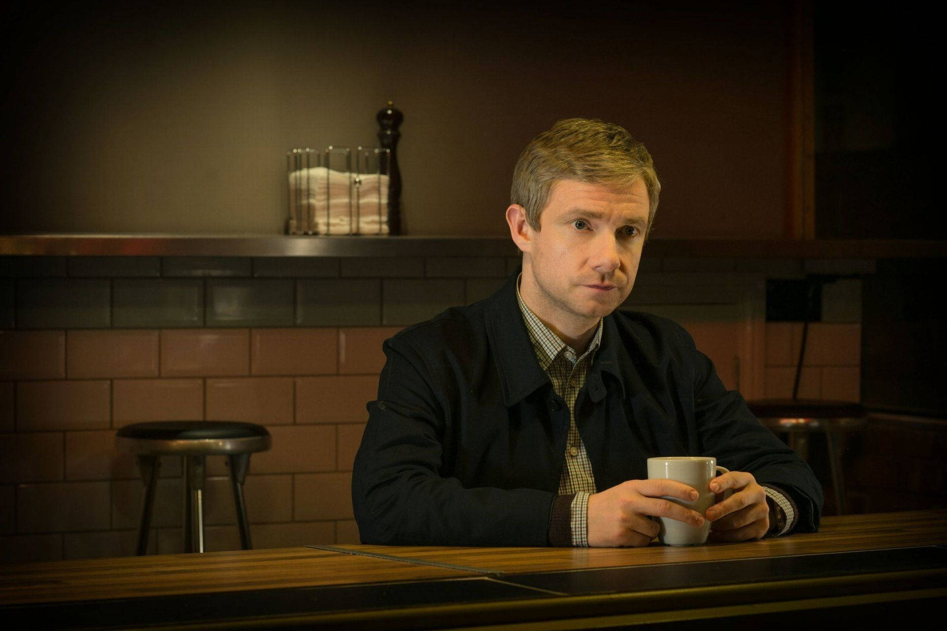 Martin Freeman 桌布 壁紙 — 一個穿著深色外套的男人坐在木吧檯前，在溫暖昏暗的燈光下，手持白色馬克杯，表情嚴肅。