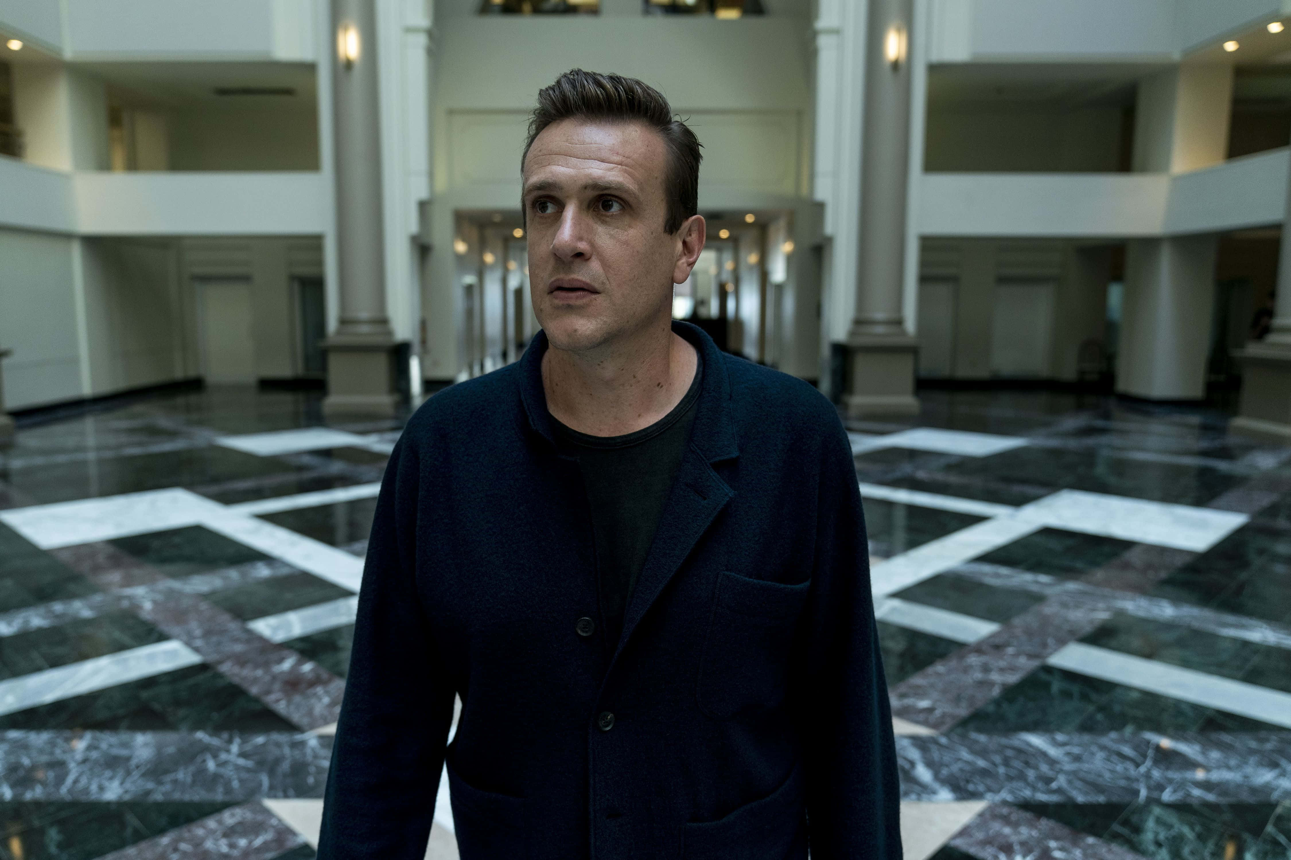 Jason Segel 桌布 壁紙 — 一個穿著深色外套的男人站在一個宏偉、空曠的大理石地板大廳，有高聳的柱子和柔和的燈光。