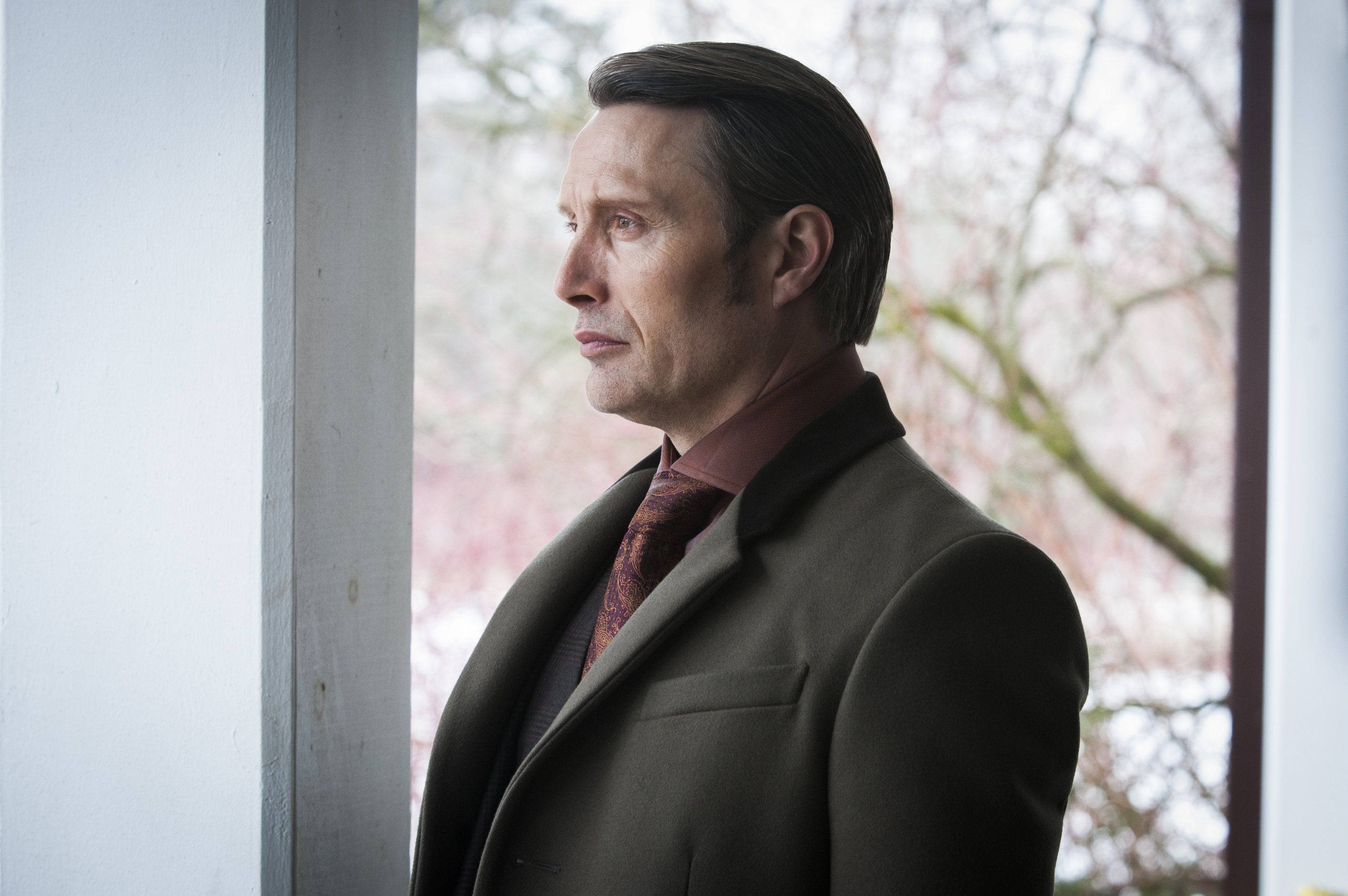 Hannibal 桌布 壁紙 — 一個穿著深色外套的男人站在窗邊，凝視著窗外光禿禿的樹木，樹上開著柔和的粉紅色花朵。