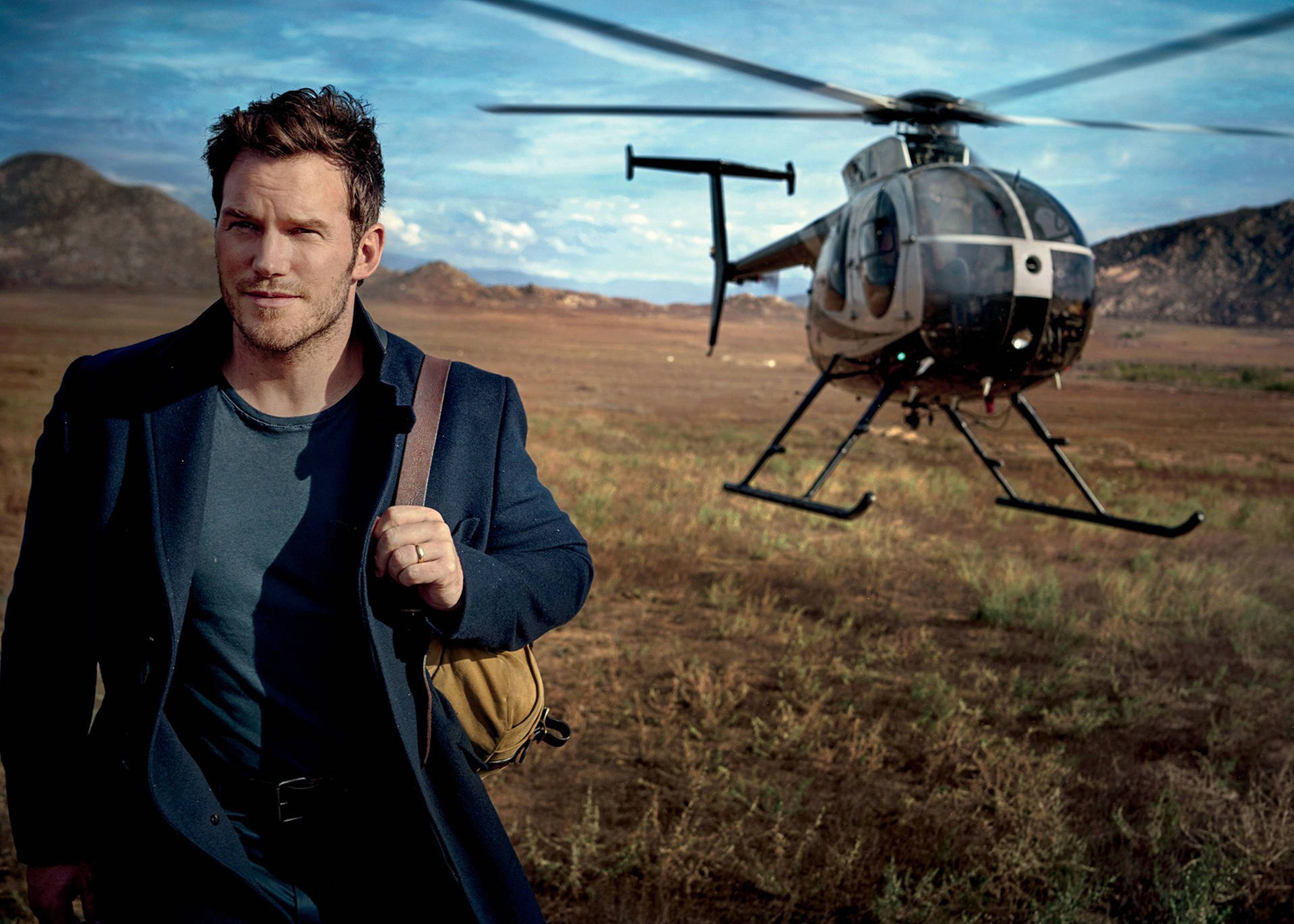 Chris Pratt 桌布 壁紙 — 一個穿著深色外套的男人自信地站在乾燥的田野上，一架直升機在他身後盤旋，背景是山脈。