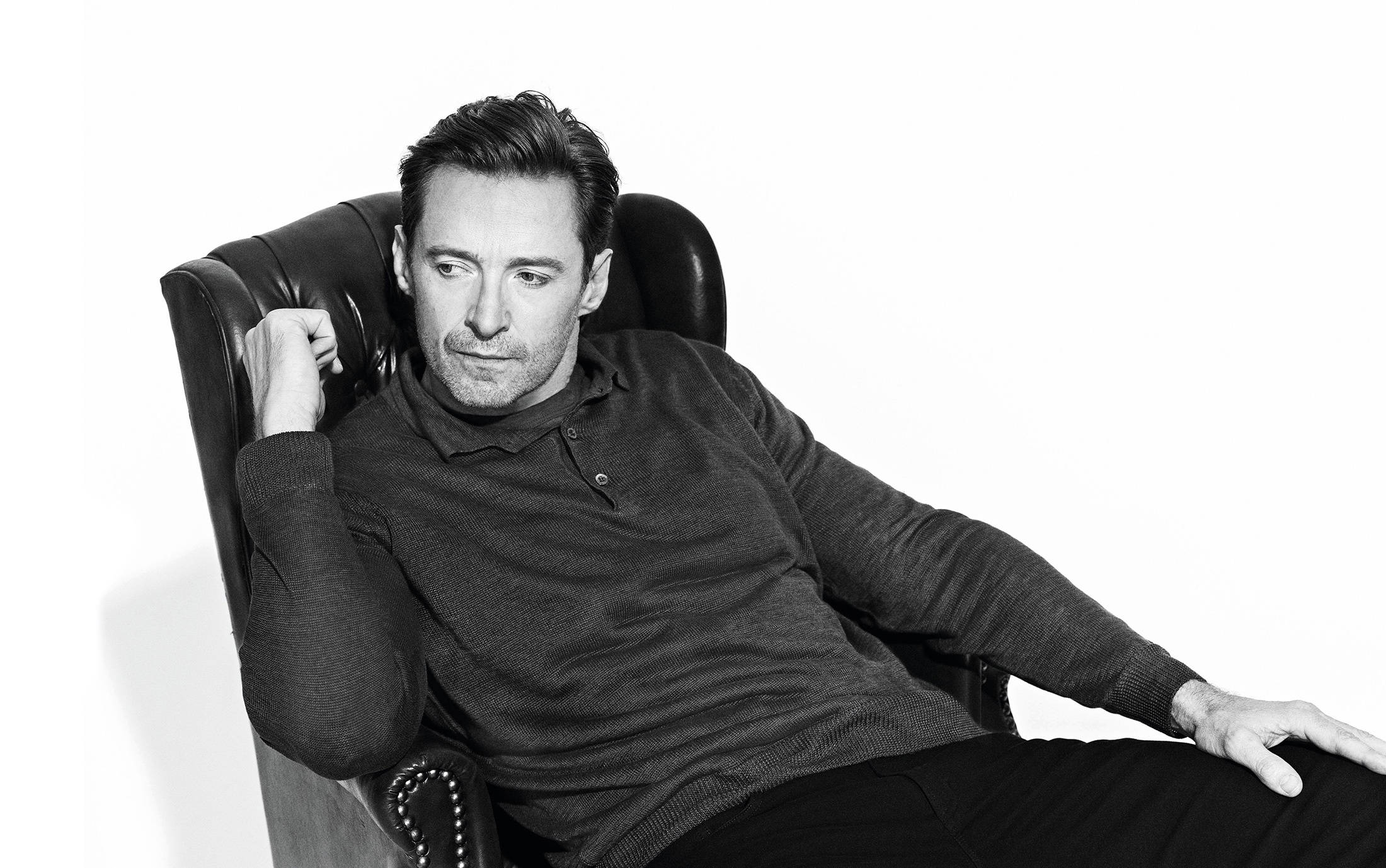 Hugh Jackman 桌布 壁紙 — 一個穿著深色毛衣的男人在白色背景前懶洋洋地坐在皮扶手椅上，一幅充滿氣氛的黑白肖像。