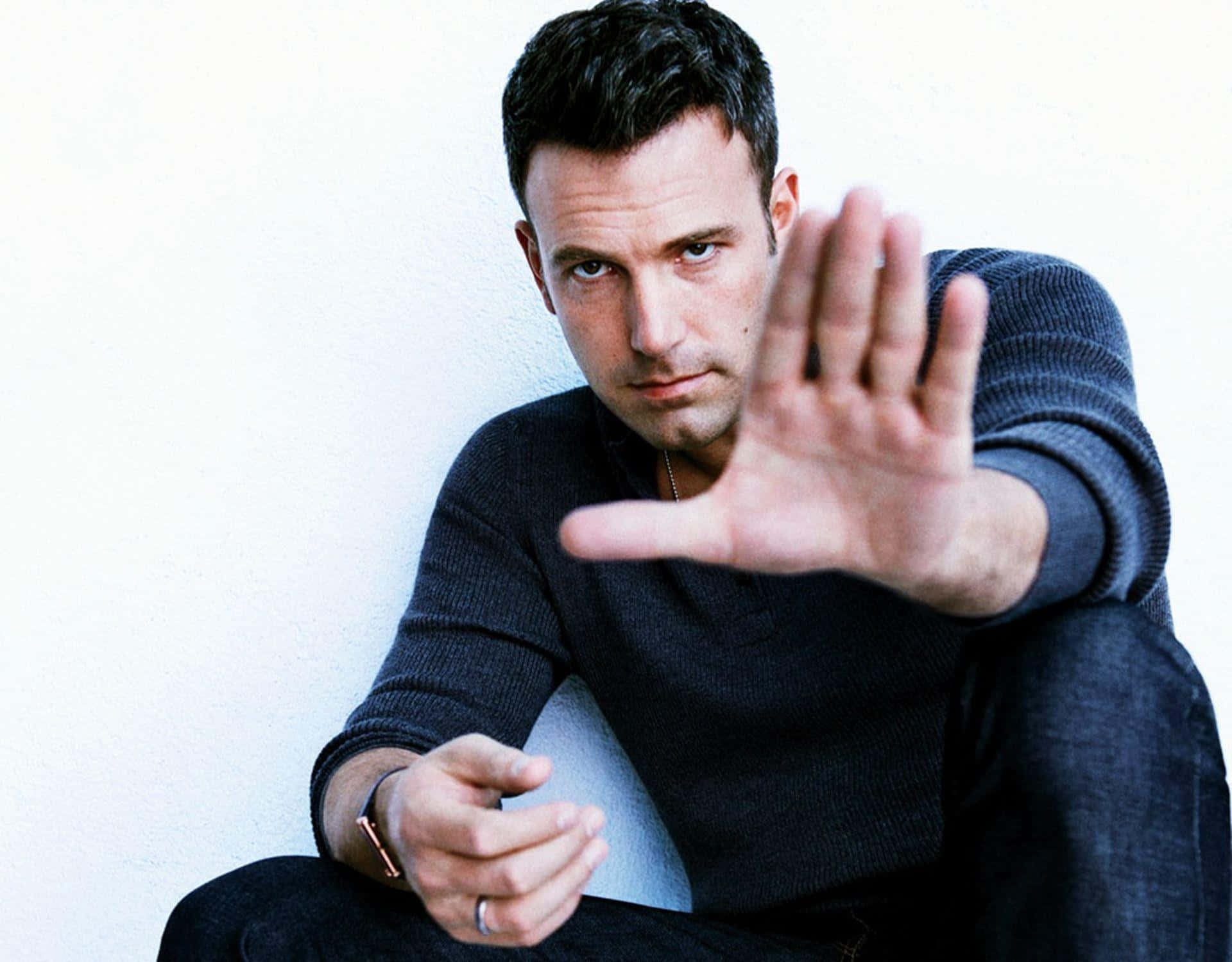 Ben Affleck 桌布 壁紙 — 一個穿著深色毛衣的男人靠著白牆坐著，表情嚴肅地向前伸出手做出停止手勢。