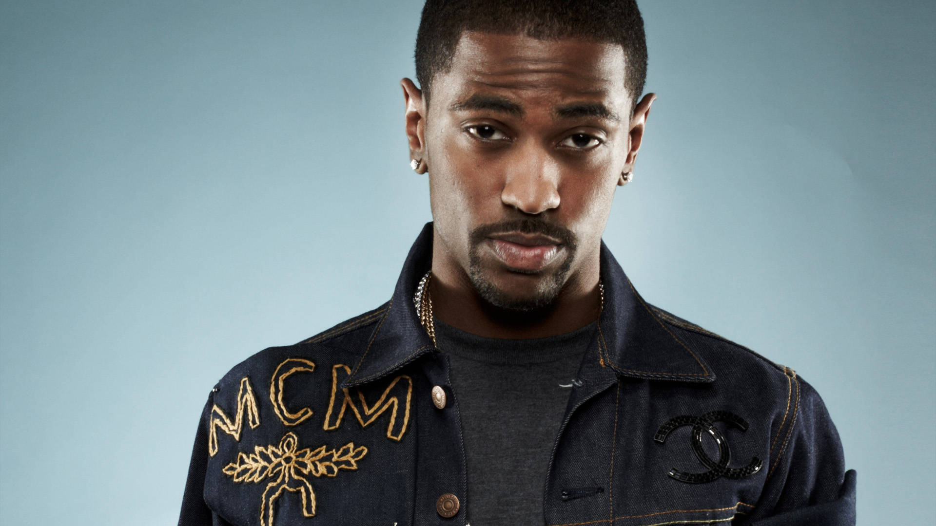 Big Sean 桌布 壁紙 — 一個穿著深色牛仔外套、帶有金色MCM刺繡和黑色Chanel標誌的男人，在柔和的藍色背景下凝視著。