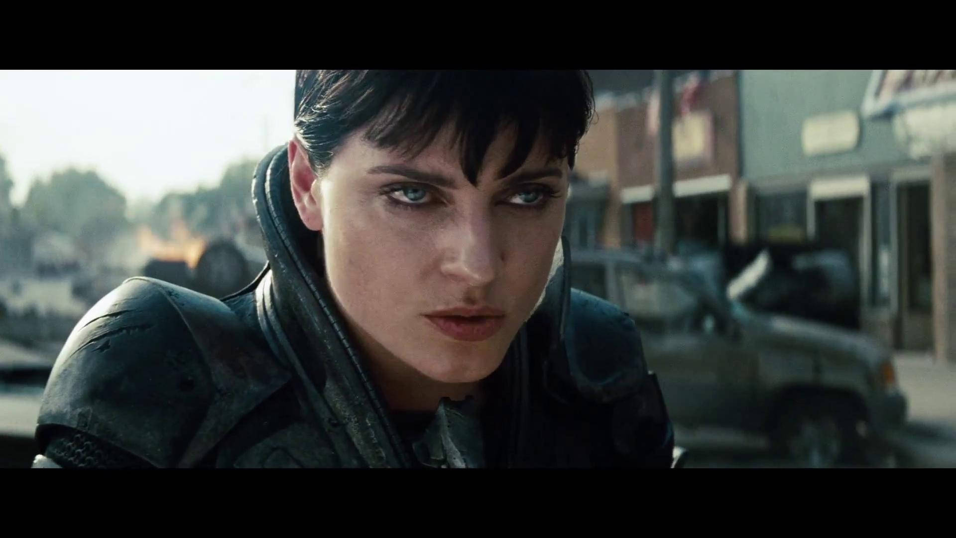 Antje Traue 桌布 壁紙 — 一個穿著深色盔甲、意志堅定的女人，在陽光普照的城市街道上凝視著，身後是模糊的汽車和建築物。