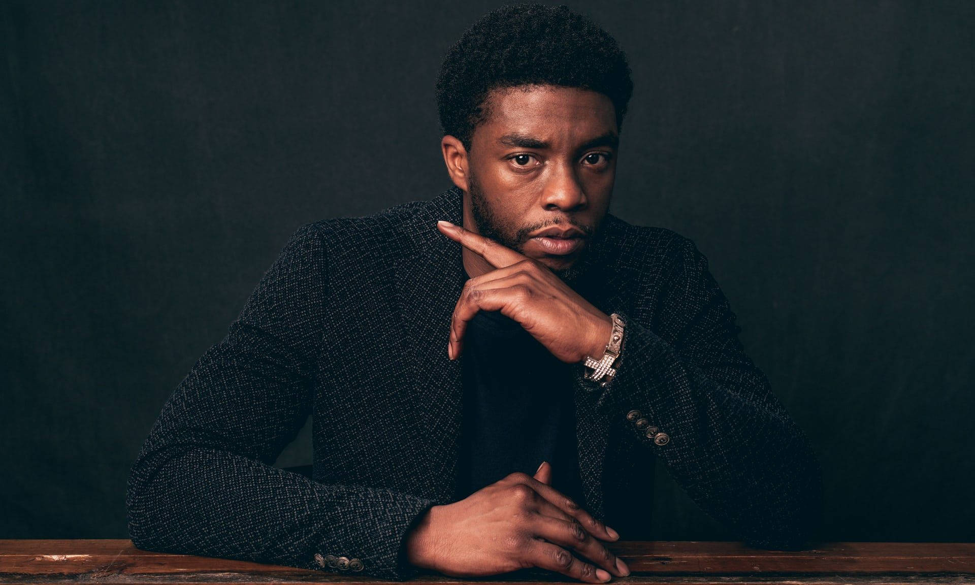Chadwick 桌布 壁紙 — 一個穿著深色紋理西裝外套的男人，在陰鬱的木炭背景下，用手托著下巴。