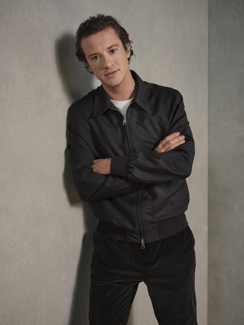 Celebrity 桌布 壁紙 — 一個穿著深色細條紋外套的男人，雙臂交叉，自信地凝視，靠在一面有紋理的灰色牆上。