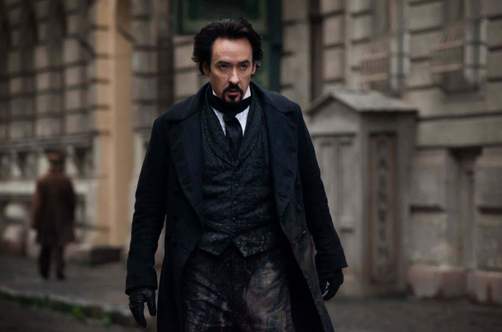 John Cusack 桌布 壁紙 — 一個穿著深色維多利亞時代外套的男人，在一條骯髒的城市街道上行走，背景有石頭建築和一個模糊的人影。