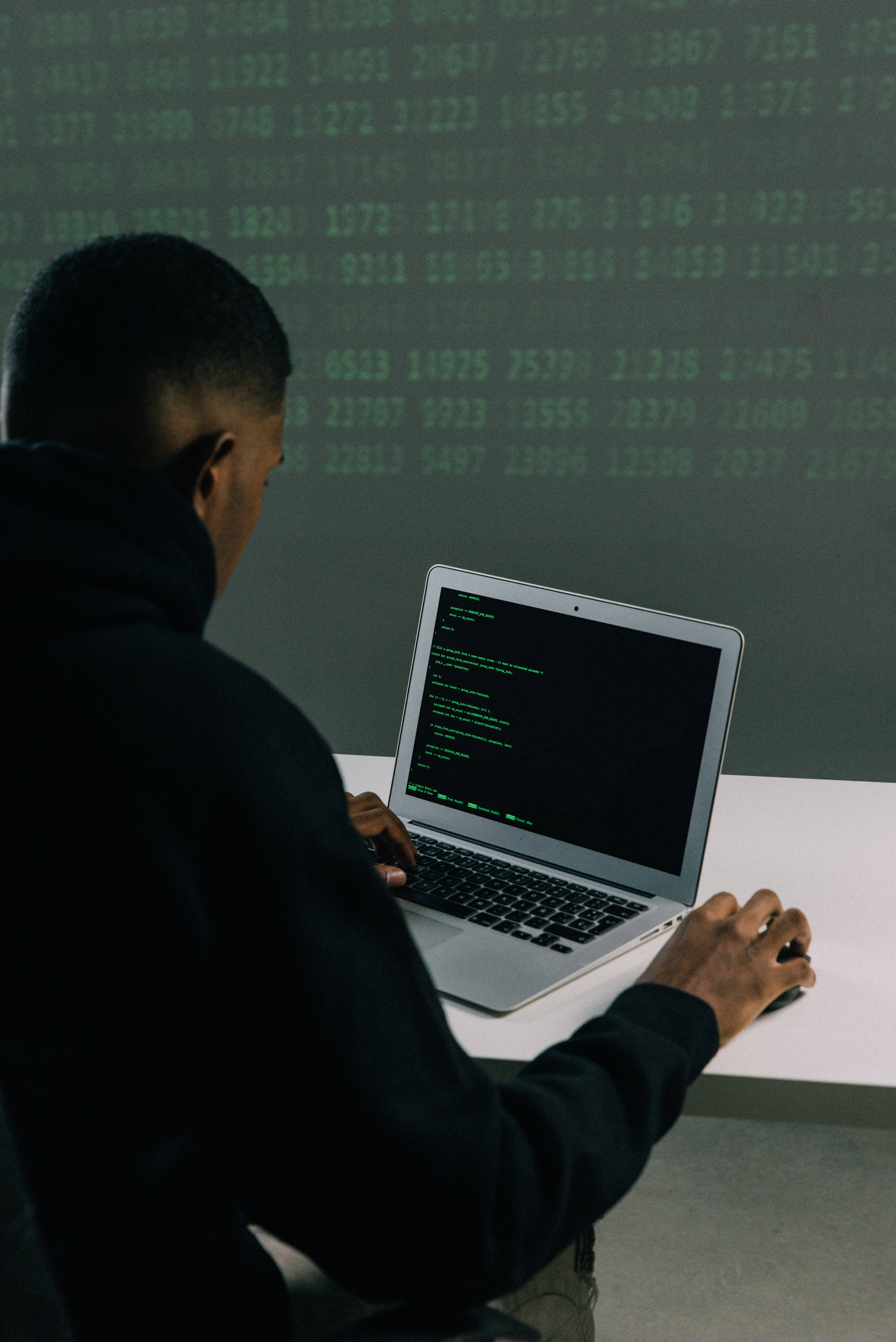 Computer Screen 桌布 壁紙 — 一個穿著深色衣服的人正在一台顯示綠色代碼的筆記型電腦上打字，背景中漂浮著發光的數字。