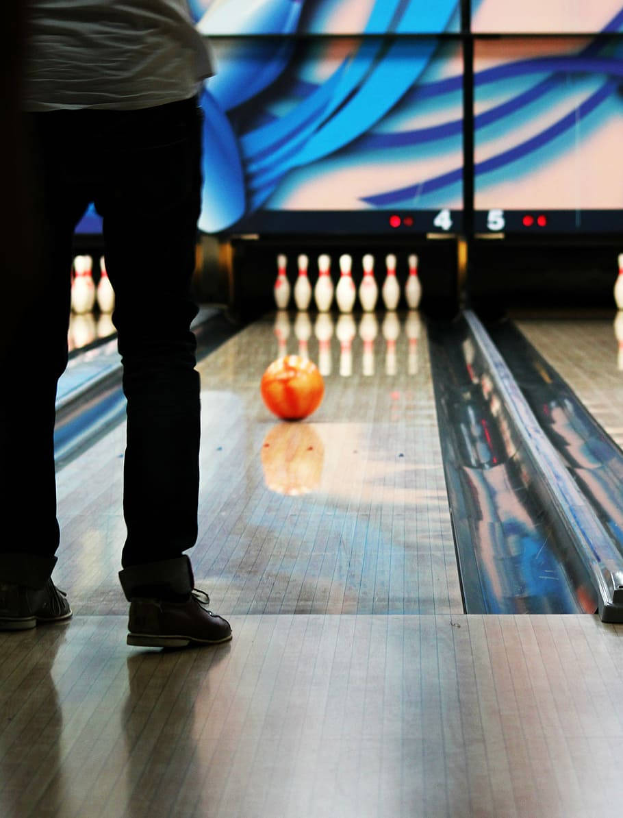bowling pin 桌布 壁紙 — 一個穿著深色褲子的人站在拋光過的保齡球道末端，一個橙色球在鮮豔的藍色燈光下滾向白色球瓶。