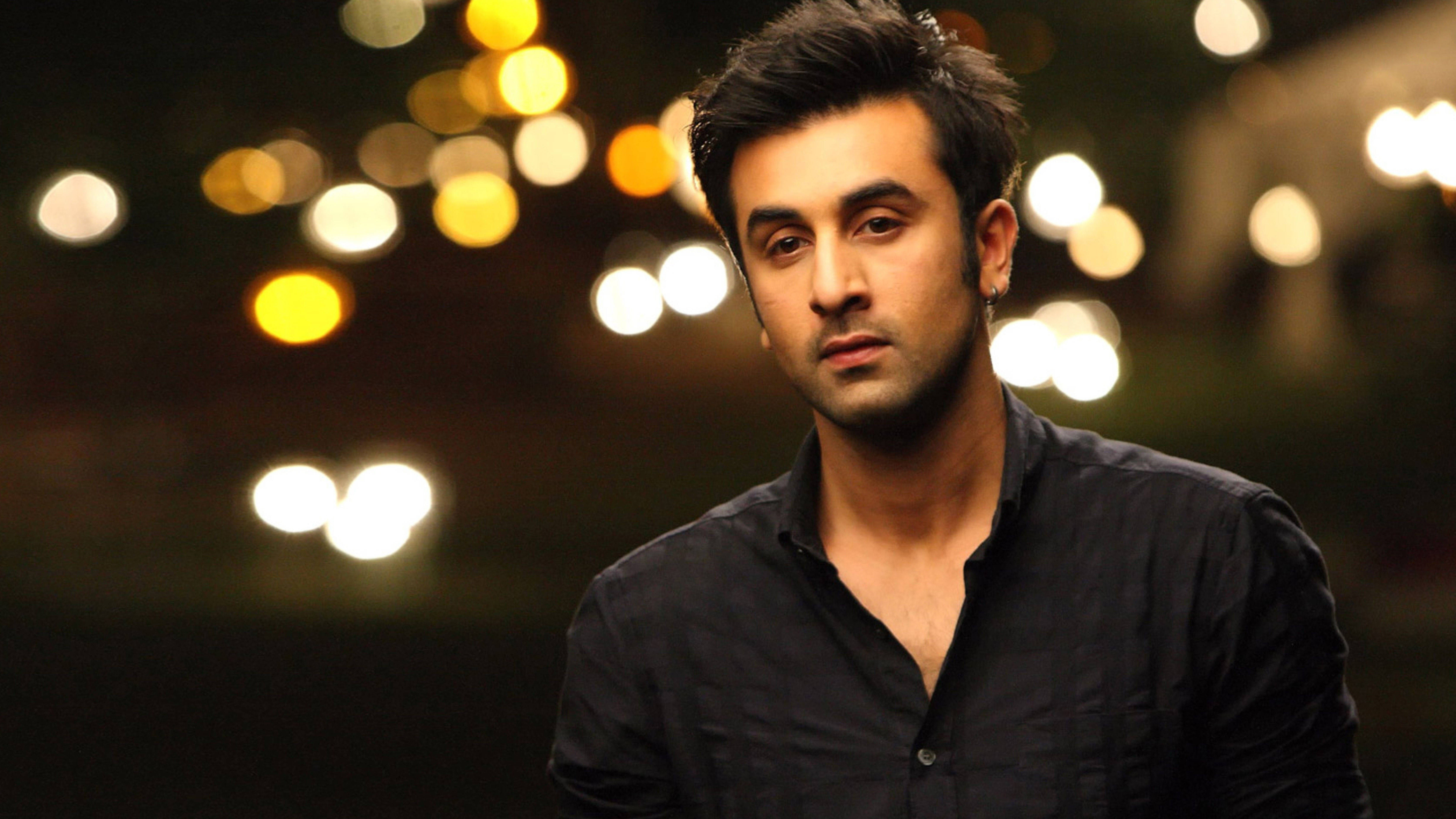 Ranbir Kapoor 桌布 壁紙 — 一個穿著深色襯衫的男人在夜晚溫暖的散景燈光背景下向前凝視。