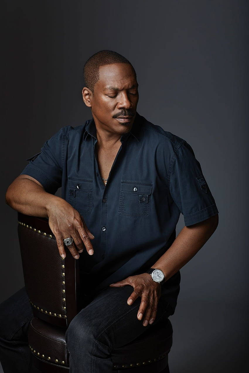 eddie murphy 桌布 壁紙 — 一個穿著深色襯衫的男人坐在棕色皮椅上，若有所思，背景是柔和的灰色。